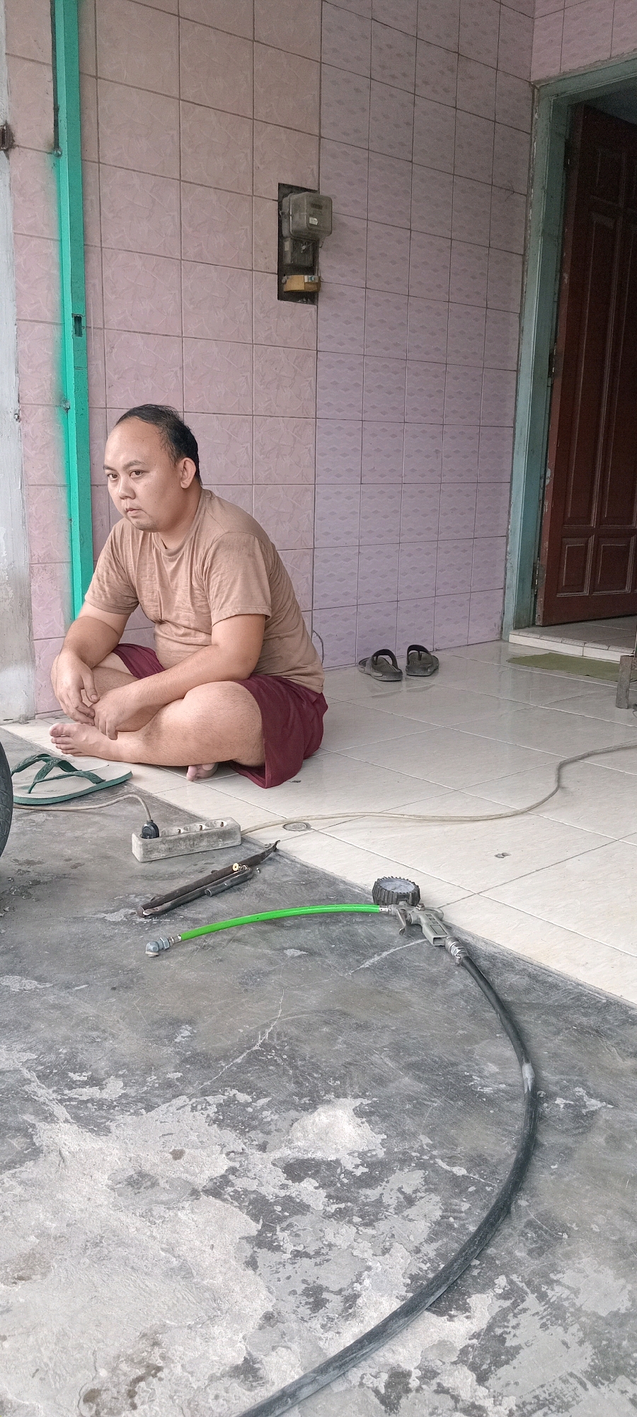In Babat, Indonesia, a man sits cross-legged on a tiled patio next to a concrete floor. He is wearing a light brown t-shirt and dark red shorts. In front of him, on the concrete floor, are various tools, including a tire pressure gauge with a green hose, a power strip with multiple outlets, and what appears to be a pair of pliers. A black hose is also visible, running along the ground. The background features a tiled wall and a wooden door. To the right of the man, a pair of dark sandals are placed on the tiled floor. The scene suggests a person engaged in or pausing from a task, possibly related to vehicle maintenance or repair, given the presence of the tire pressure gauge and hoses.