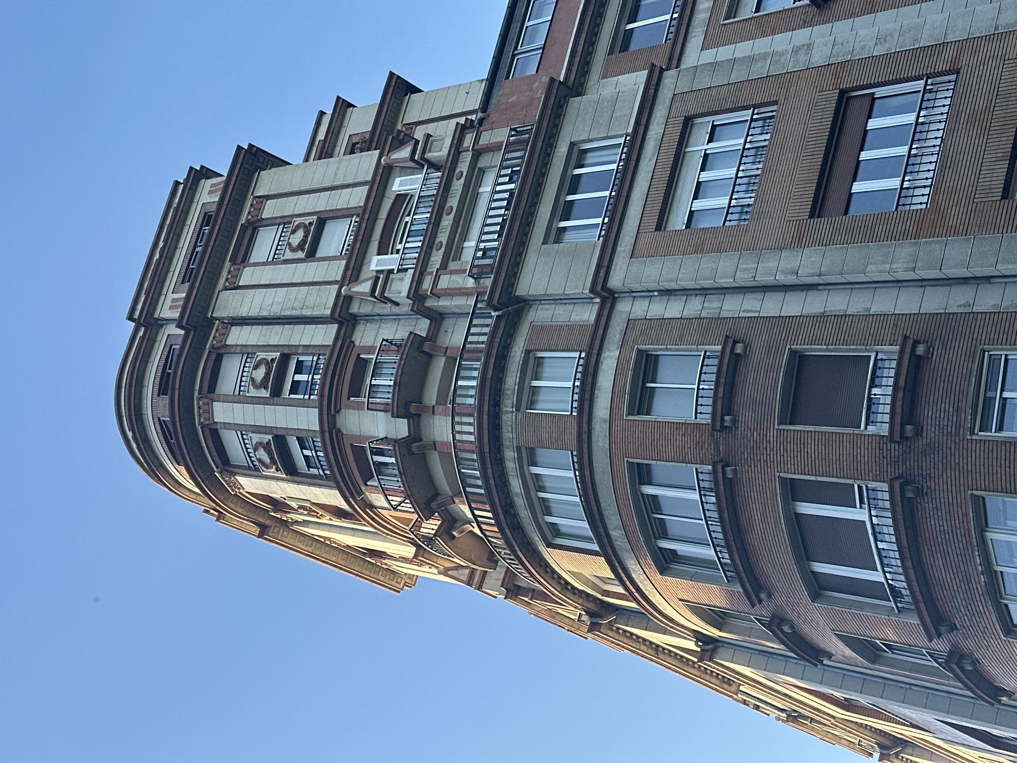 The image shows a low-angle, upward view of a multi-story building with a curved facade against a clear blue sky. The building is constructed with brick and light-colored stone elements, featuring decorative architectural details such as columns, pilasters, and balconies with wrought-iron railings. Many windows are visible, some with internal blinds drawn, reflecting the bright sky. The architectural style suggests it is an older building, possibly from the early to mid-20th century. This building is located in Oviedo, Spain, specifically on Calle Uría, a prominent street known for its elegant architecture. There are no people visible in the image.