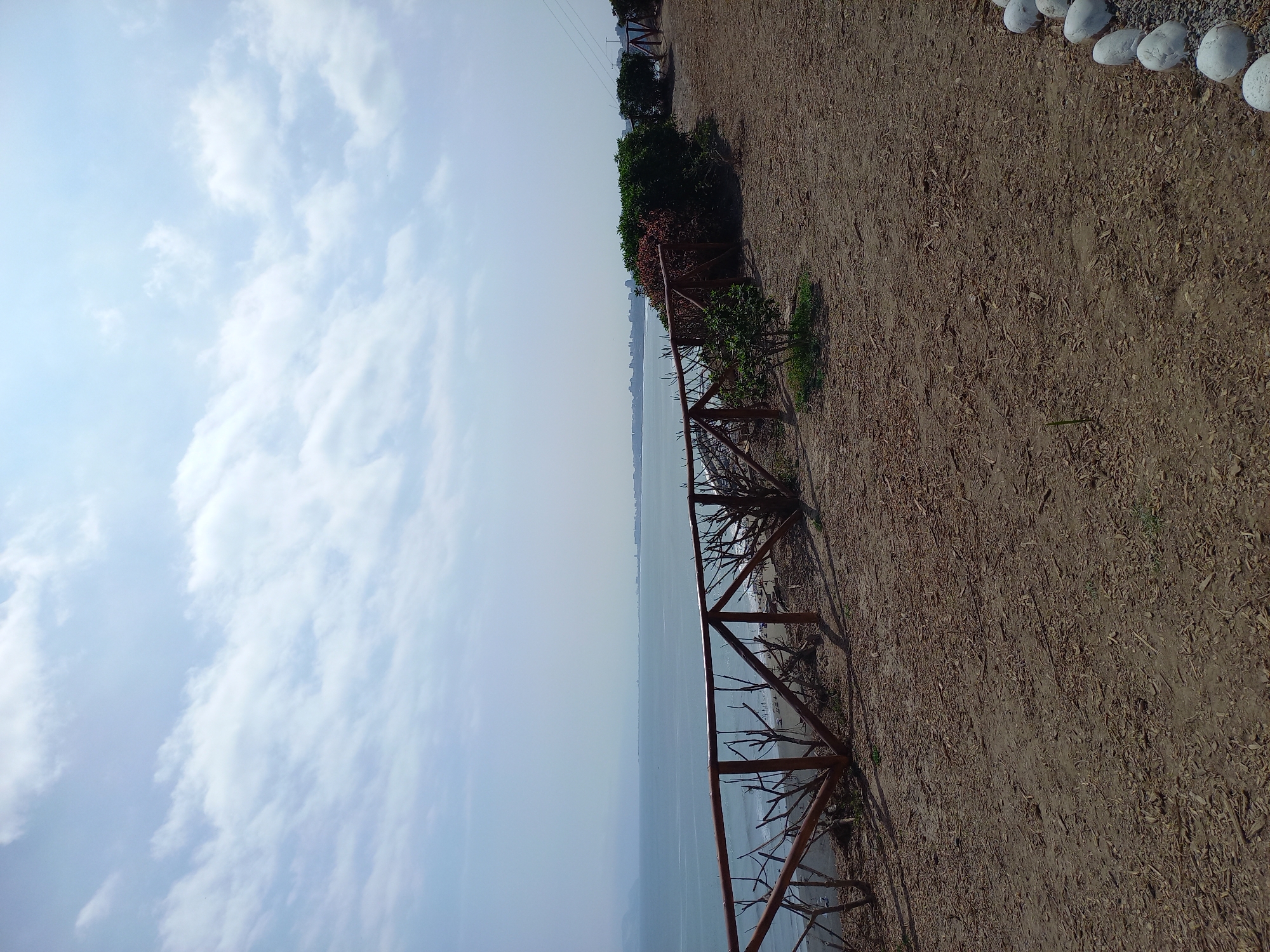 The image shows a scenic view of the ocean from a bluff. The scene is dominated by the vast expanse of the ocean under a bright, cloudy sky. A dirt path runs along the top of the bluff, lined with a rustic wooden fence made of branches. Beyond the fence, there are some green bushes and trees. In the distance, a city skyline is visible, hinting at an urban setting on the coast. The time of day appears to be daytime, with ample natural light. The weather seems pleasant, with scattered clouds providing some shade against the sun. The location is identified as Chorrillos, Peru, suggesting a coastal region with a city nearby. There are no people or specific activities depicted in the image; it's a tranquil, observational shot of the landscape. The overall mood is peaceful and serene, capturing the beauty of the natural environment meeting the cityscape.
