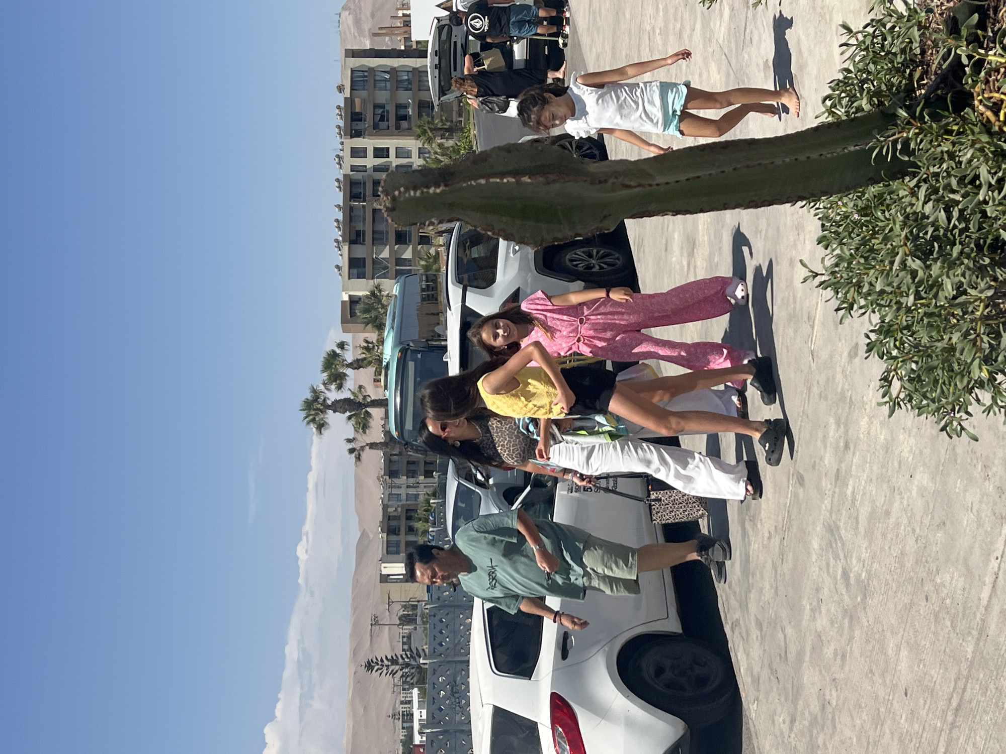 The image shows a group of people standing on a paved area with several vehicles parked nearby. In the background, there are buildings and a sandy landscape. The sky is clear and blue. A large cactus is visible in the foreground. The people in the group appear to be interacting with each other. It is unknown the exact location in Arica, Chile.
