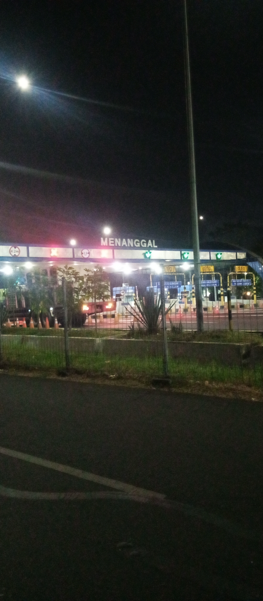 The image depicts the Menanggal toll gate at night in Surabaya, Indonesia. A brightly lit toll plaza is visible with signs indicating lane information, including vehicle height restrictions and directions. There are vehicles at the toll booths, some with their headlights on, and traffic cones are strategically placed. The foreground shows a dark road with lane markings, and a fence separates the road from the toll plaza area. The background is dark, with a bright light source in the upper left corner, possibly a street lamp. The overall scene captures a functional view of a toll collection point during nighttime hours.