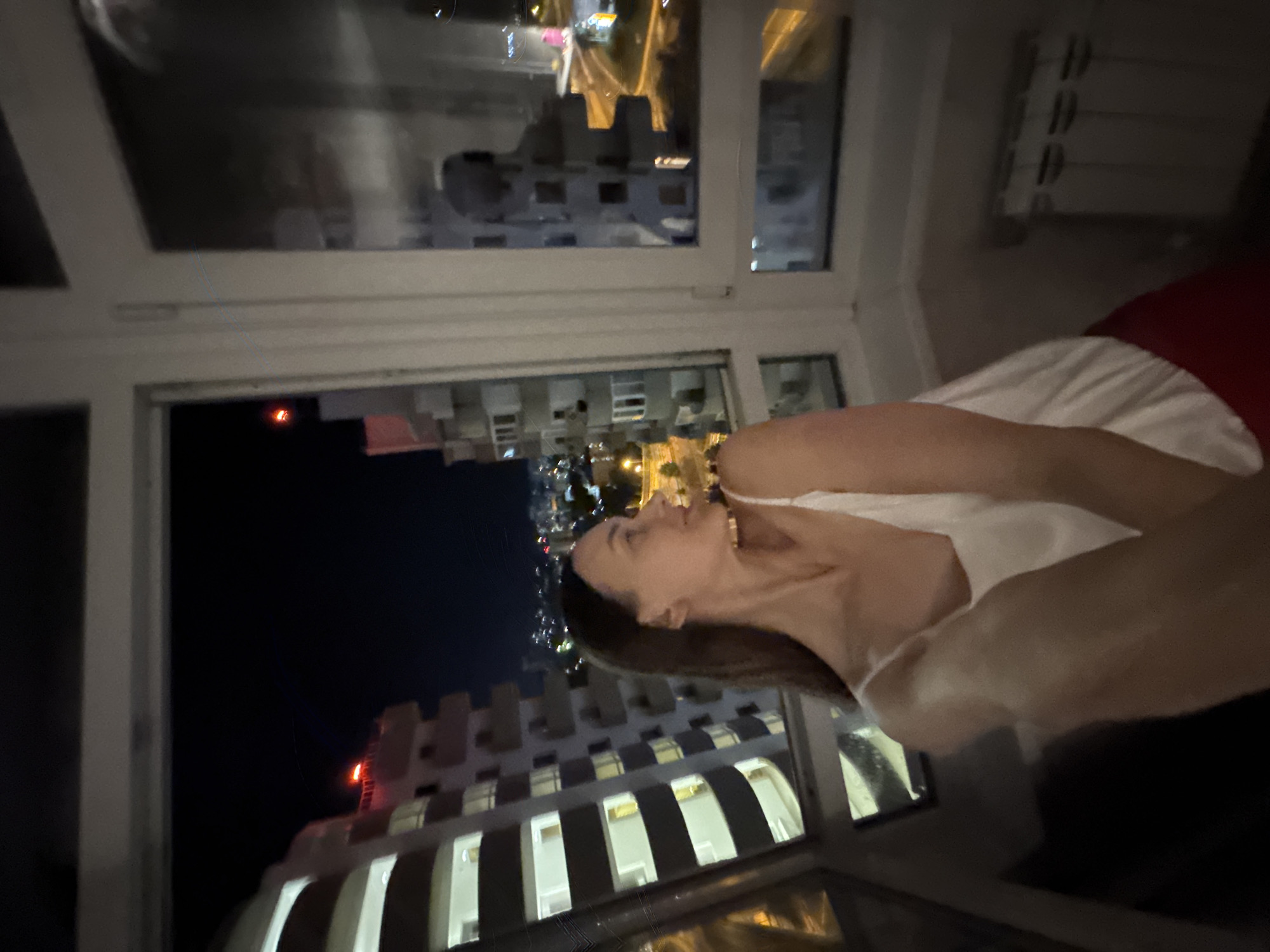 A young woman, with long dark hair, is captured in a low-angle, medium shot taken at night from inside an apartment. She is wearing a white camisole and red bottoms. The woman is looking to her right, away from the camera, with a slight smile. She is positioned in front of a large window that offers a view of a city at night. In the foreground, partially visible, are the woman's arms and shoulders. The background shows a cityscape with illuminated buildings and roads. The scene suggests a residential setting, likely an apartment balcony or room with a panoramic view. The lighting is dim, with the primary light source appearing to be the city lights and the interior apartment lighting. The overall impression is one of a private, contemplative moment enjoyed with a city view. The crops indicate the location is in Sochi, Russia.