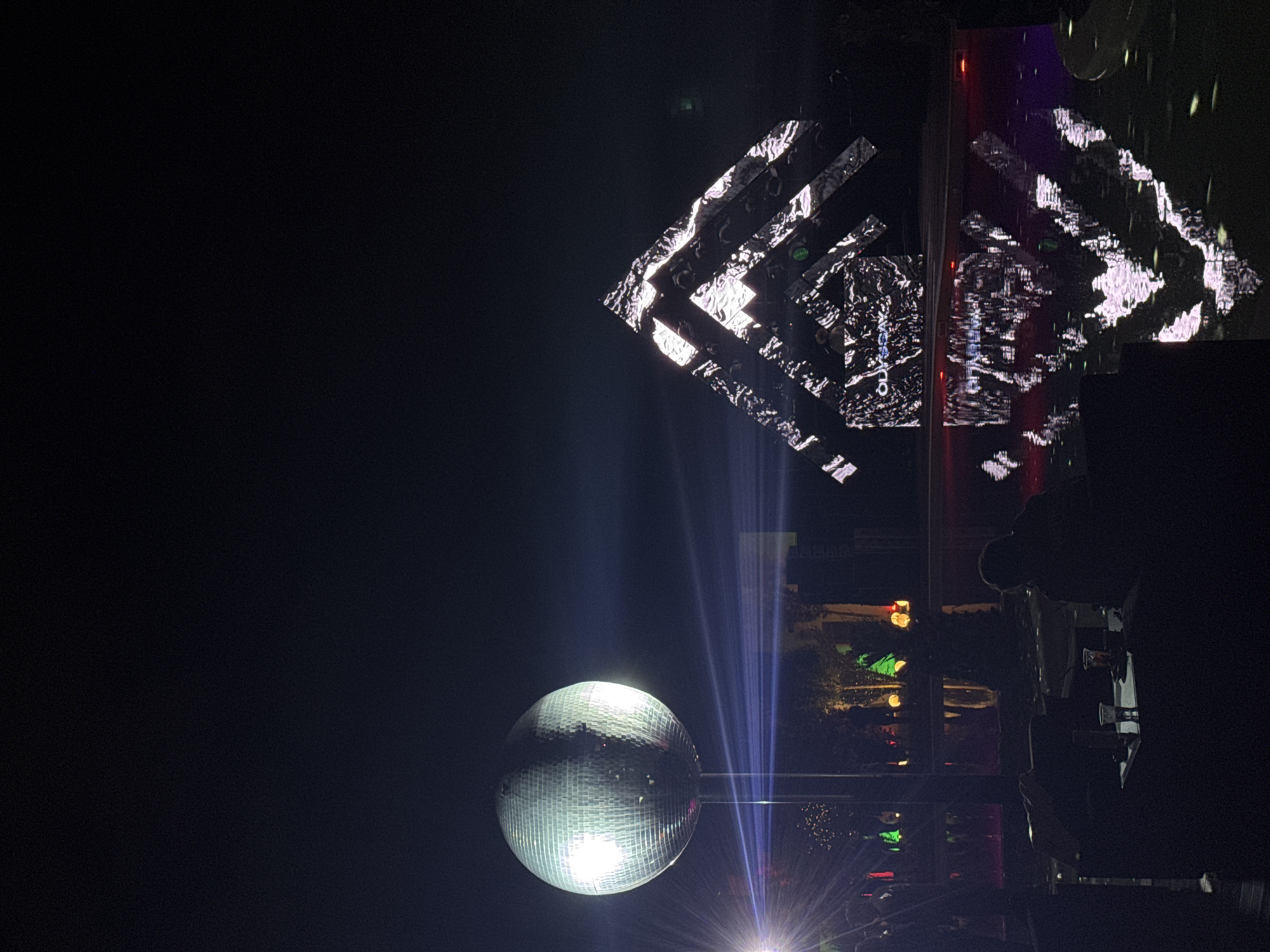 The image shows a nighttime scene, possibly an event or concert, likely in Parakar, Armenia based on the visible "ARARATPARK" sign. In the foreground, a reflective disco ball is illuminated by spotlights, creating bright rays of light shooting upwards. Above and slightly to the right of the disco ball, a complex structure of rectangular panels displays a geometric pattern in black and white. The scene appears to be crowded with people, though they are mostly silhouettes in the dim lighting. The background contains blurred lights and vegetation, indicating an outdoor setting within a park or public space.