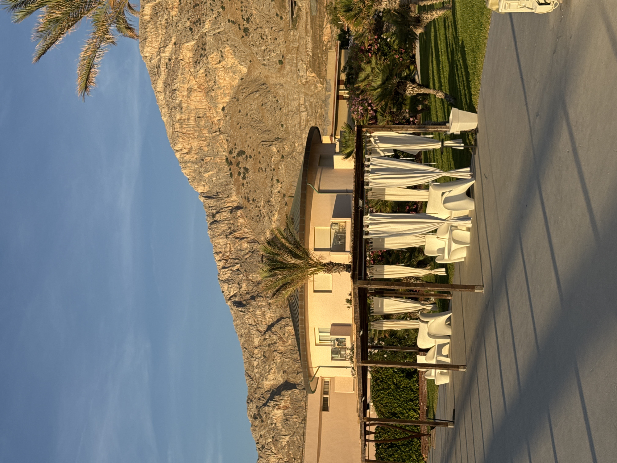The image depicts a scene in Isola delle Femmine, Italy, featuring a resort-like setting with a mountainous backdrop. In the foreground, a paved walkway is visible with long shadows cast by a wooden structure supporting white curtains. This structure appears to be an outdoor seating area, furnished with white, modern-looking chairs. Beyond this seating area, a pale pink building with rounded architectural elements and visible windows is situated. A palm tree stands in front of this building. The background is dominated by a large, rugged, sun-drenched mountain range. Lush green grass and tropical vegetation, including palm trees and flowering plants, surround the building and seating area. The sky is a clear blue with a few faint wisps of clouds. The lighting suggests it is either early morning or late afternoon, with the sun casting a warm glow on the mountain and illuminating the scene. There are no people visible in the image.