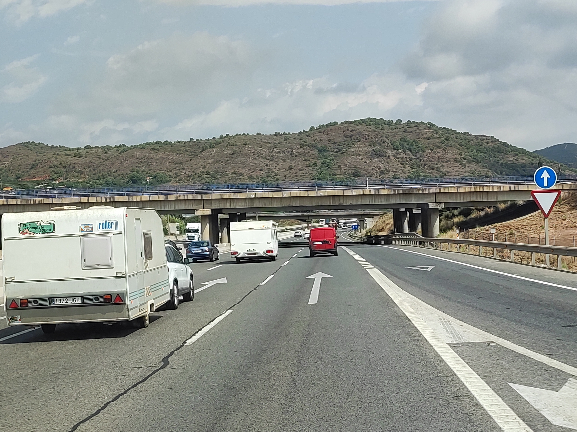 The image captures a highway scene with several vehicles, including a white caravan being towed by a car, a red van, and other cars and trucks in the distance. A bridge passes overhead, and a sign with an upward arrow is visible on the right side of the road. The landscape is characterized by dry, hilly terrain with sparse vegetation, typical of the Mediterranean climate. The sky is cloudy. The caravan on the left has a license plate that reads "1872 JGH". This scene is likely situated on a highway near Sagunto, Spain, given the style of the road signs and the surrounding landscape.