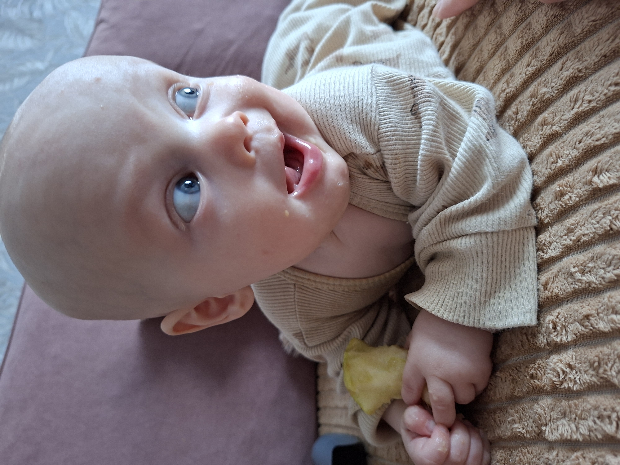 A close-up, high-angle shot depicts a baby lying down in an indoor setting in Karsha, Russia. The baby has a bald head, fair skin, and wide, bright blue eyes that gaze upwards and slightly to the side. Its mouth is open, revealing a pink tongue, and there is visible food residue and saliva around its lips, indicating it is eating or has recently eaten.
The baby is dressed in a light beige, ribbed long-sleeved top, which appears to have a few small smudges or stains. Its small hands are gripping a light yellow piece of food, resembling an apple slice. The baby's expression conveys a sense of engagement and curiosity. The immediate background features a textured brown surface, possibly a blanket or soft furnishing, while behind the baby's head is a muted purple or pink cushion or sofa. There is no visible text in the image.