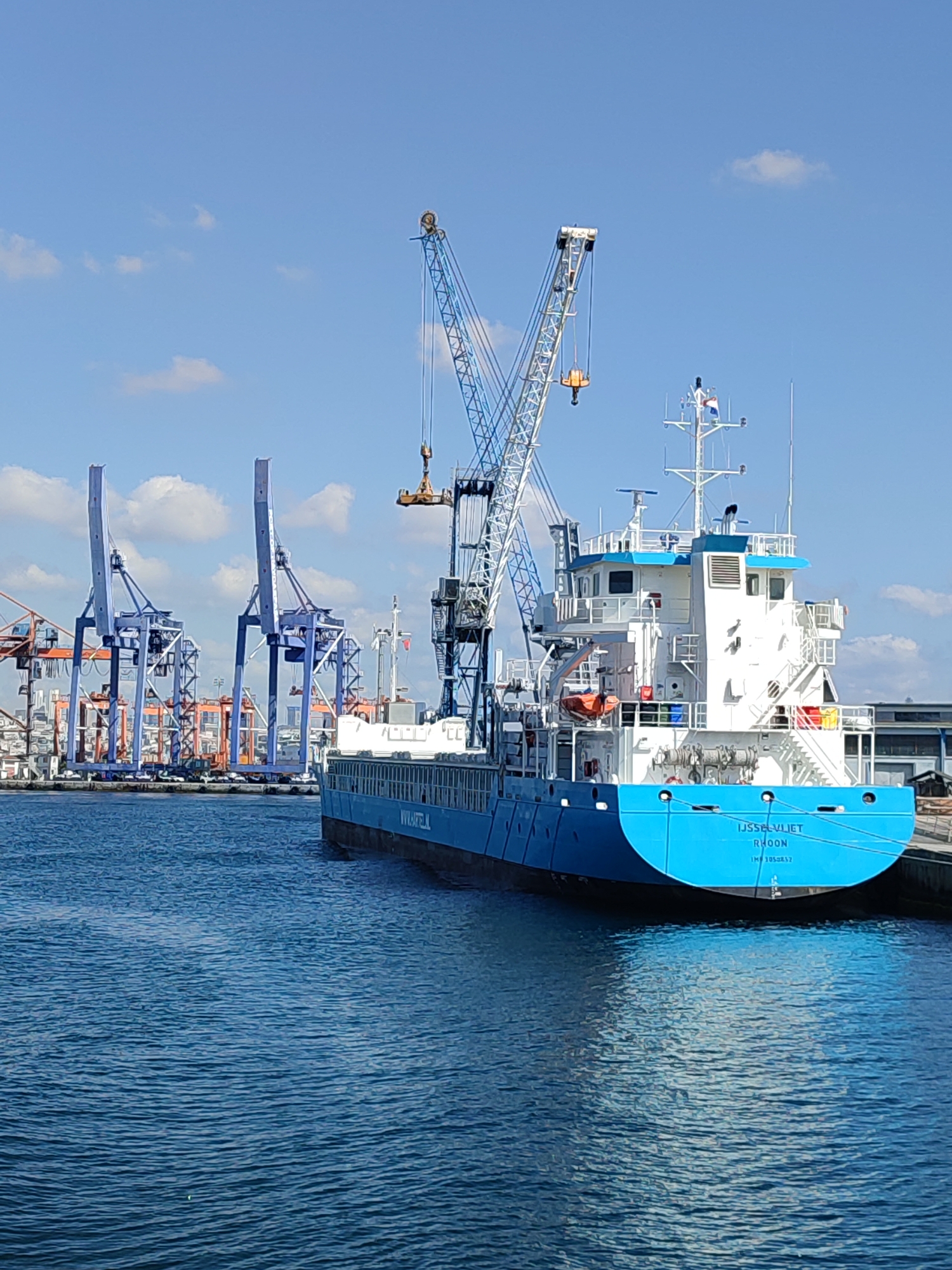 In Istanbul, Turkey, a cargo ship named "IJsselviet" is docked at a port on a clear, sunny day. The ship, painted blue and white, is a significant subject in the foreground, positioned close to the viewer. It appears to be either loading or unloading cargo, as several large cranes are visible in the background, some actively engaged in the operation. The water is a deep blue and shows gentle ripples, reflecting the bright sky.
In the background, a series of large, industrial cranes, primarily blue and orange, dominate the port landscape. These cranes suggest an active shipping and logistics hub. The ship itself has visible markings, including "IJsselviet" and "RHOON" on its stern, along with an IMO number. The ship's superstructure is white, with multiple decks and navigational equipment. There are no people clearly visible on the deck of the ship or in the immediate vicinity. The overall scene depicts a busy commercial port environment in Istanbul.