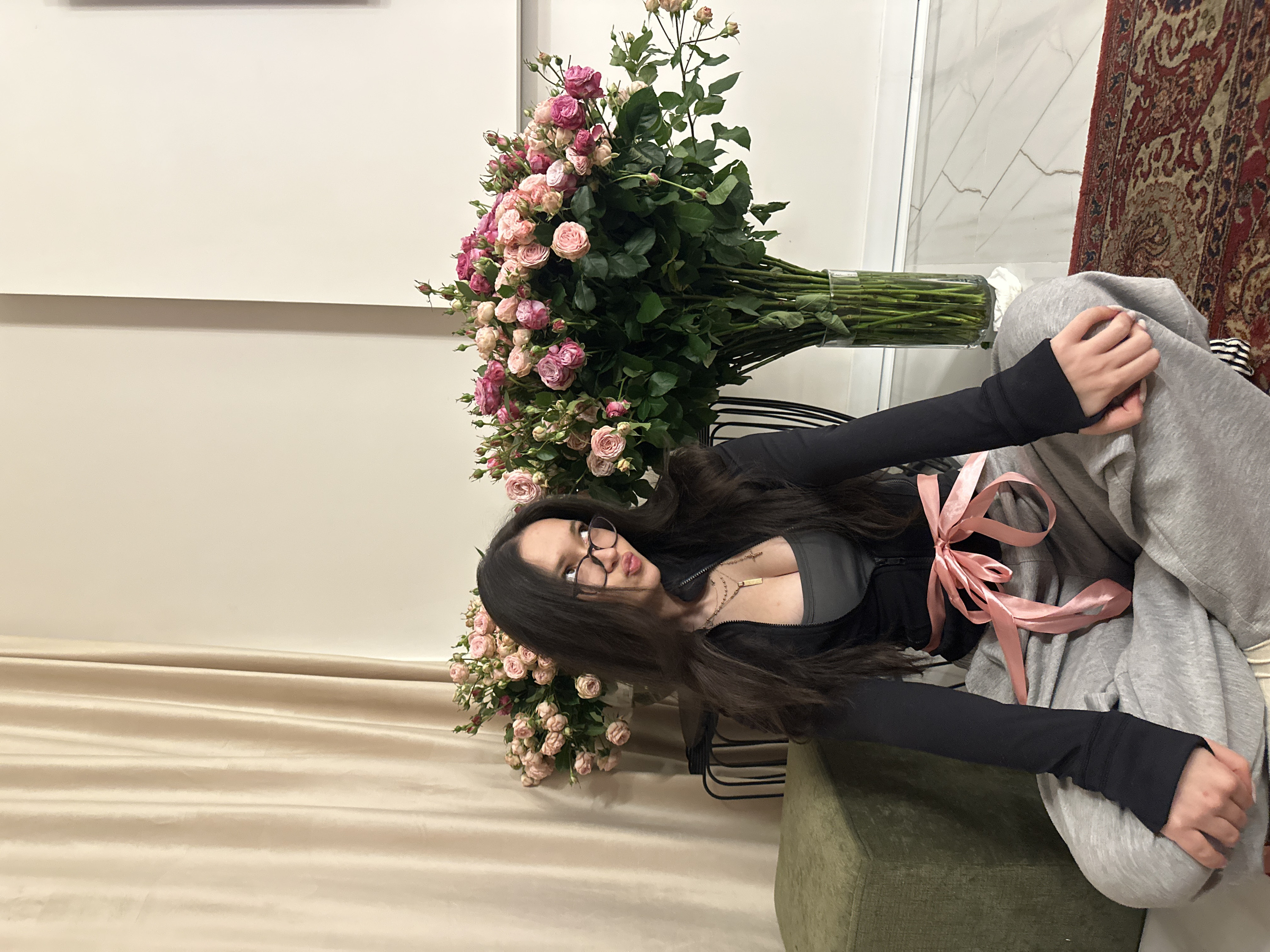 The image displays a young woman seated next to a large bouquet of flowers in an indoor setting. The image is oriented sideways, with the woman and flowers appearing to be on their sides.
The woman has long, dark hair that cascades around her shoulders and chest. She is wearing eyeglasses with dark frames. Her top consists of a dark gray or black long-sleeved athletic-style jacket, partially unzipped, revealing a dark green or gray top underneath. Around her midsection, a light pink satin ribbon is tied in a bow, adding a decorative element to her outfit. She is also wearing a layered necklace with at least two chains, one of which has a rectangular pendant. Her lower body is covered by light gray sweatpants or similarly comfortable trousers. One hand, with light-colored fingernails, is visible, resting on her leg. Her expression is somewhat subtle, with her lips slightly parted.
To her right (when viewed upright), an abundant bouquet of pink roses, varying in shades from delicate light pink to deeper rose, is arranged in a clear glass vase. The roses are still largely in bud form, with many smaller buds among the more open blooms. The stems and green foliage are prominently visible in the vase. The flowers appear to be resting on a surface, possibly a low table or bench.
The background features a light-colored, possibly off-white or cream-colored, wall behind the flowers and the woman. To the far right, there is a portion of a red and cream patterned rug or carpet visible, along with what appears to be a tiled wall with a subtle marbled or crackled texture. Below the woman, a muted olive-green cushion or part of a chair is visible. To the left, a light beige or cream-colored curtain or drapery with soft folds adds texture to the lower left quadrant of the image.
The lighting appears to be indoor ambient light, creating soft shadows and highlighting the details of the flowers and the woman's features. There is no visible text in the image.
