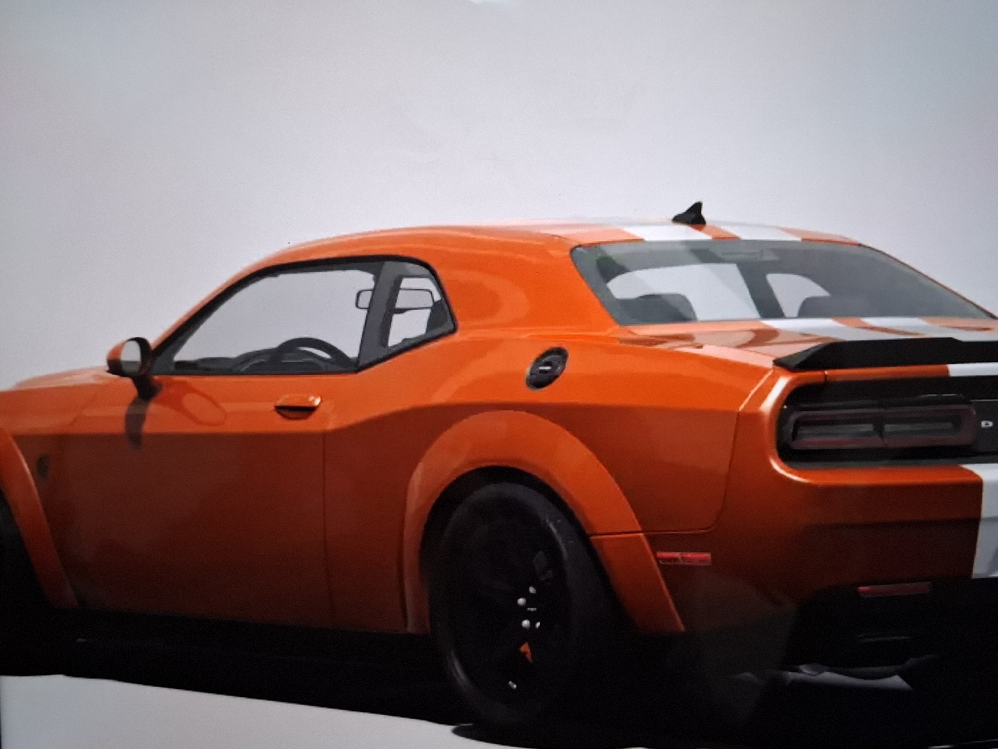 An orange Dodge Challenger with white racing stripes is parked on a white surface, possibly a display floor or studio. The car is viewed from the rear-quarter angle, showcasing its wide fenders, black wheels, and prominent rear spoiler. The background is a plain, light gray, indicating an indoor setting. There are no people or other objects visible in the scene. The image does not contain any elements that would suggest a location in the city of Argun, Russia.