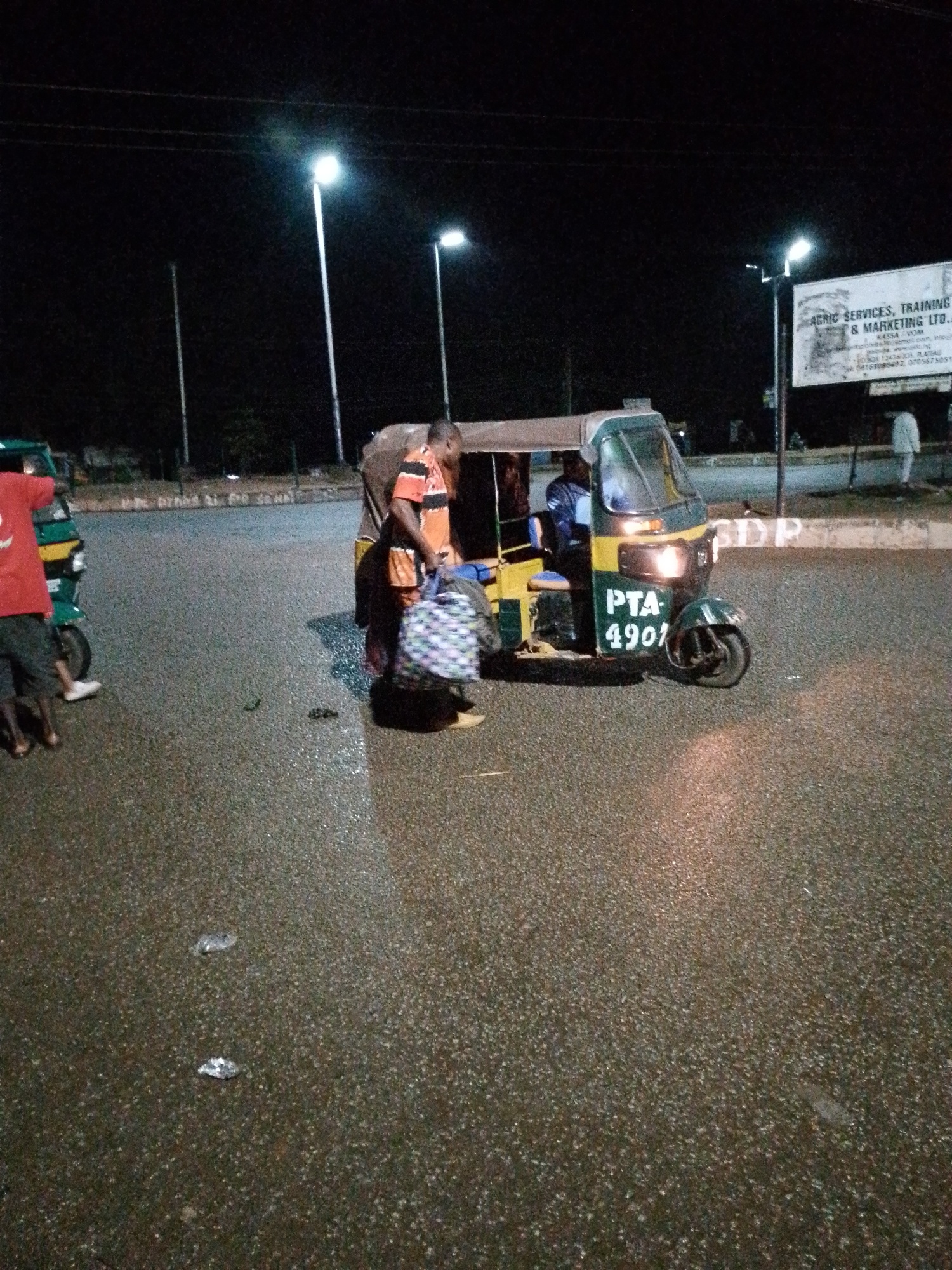 At night in Bukuru, Nigeria, a yellow and green auto-rickshaw with the license plate "PTA-4901" is parked on a wet asphalt road. The auto-rickshaw's headlights are illuminated. A person wearing a red shirt and dark shorts stands to the left of the auto-rickshaw, facing away from the camera. Another person, wearing an orange and black striped shirt, stands next to the auto-rickshaw on the right, holding a patterned bag. A white sign is visible in the background, with text that is partially legible. Streetlights illuminate the scene, reflecting off the wet road surface. The environment appears to be an intersection or a busy road.