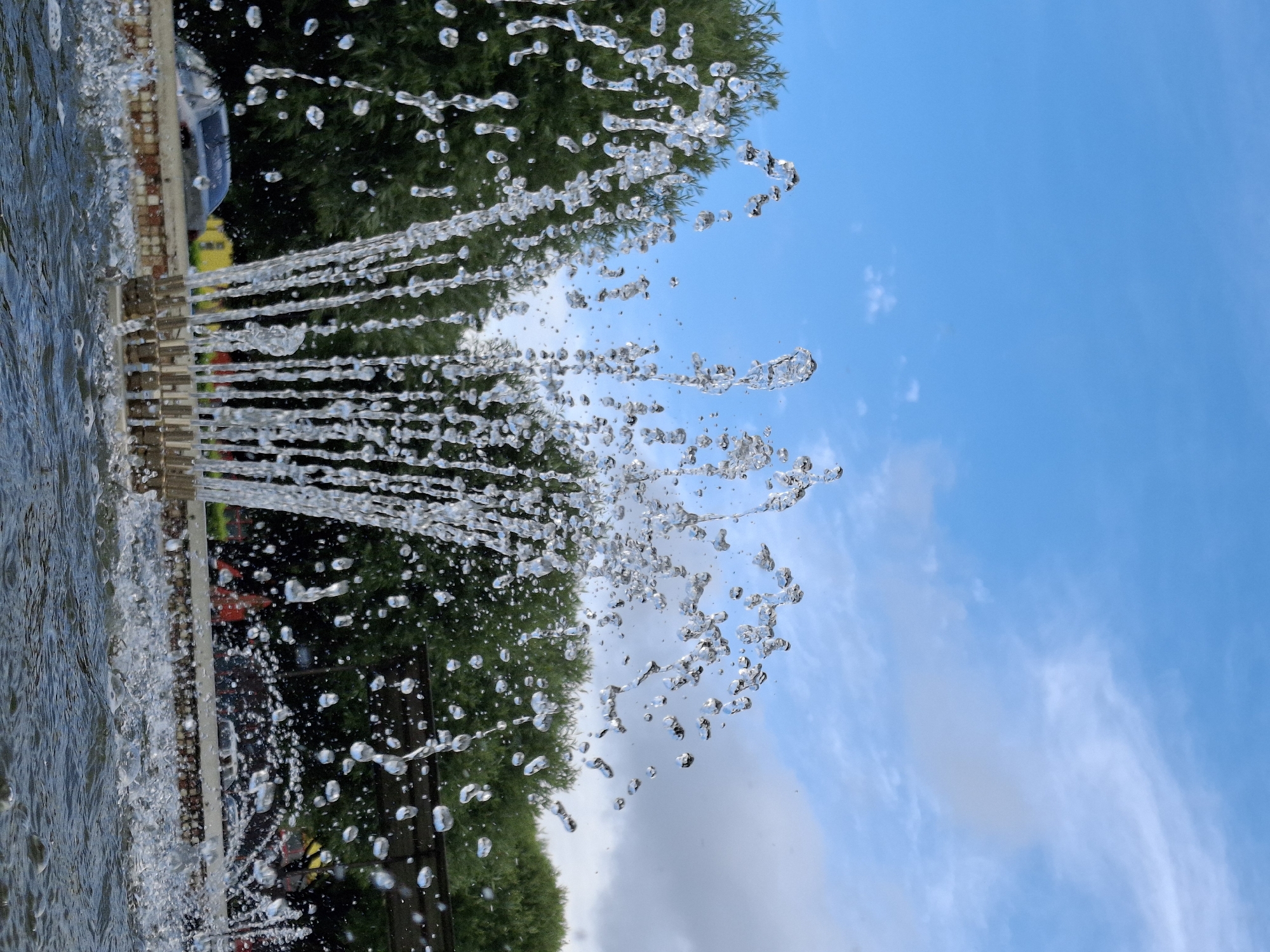 In a close-up, low-angle shot, a fountain sprays water upwards against a backdrop of a bright blue sky with scattered white clouds. Lush green trees are visible behind the fountain, with a hint of a dark structure and a glimpse of a silver car and yellow and red elements in the distance, suggesting an outdoor urban setting. The water droplets are frozen in mid-air, creating a dynamic display of liquid sculpture. The image is taken from the perspective of someone standing near the water's edge, with ripples and splashes visible in the foreground. This scene is located in Chaikovsky, Russia.