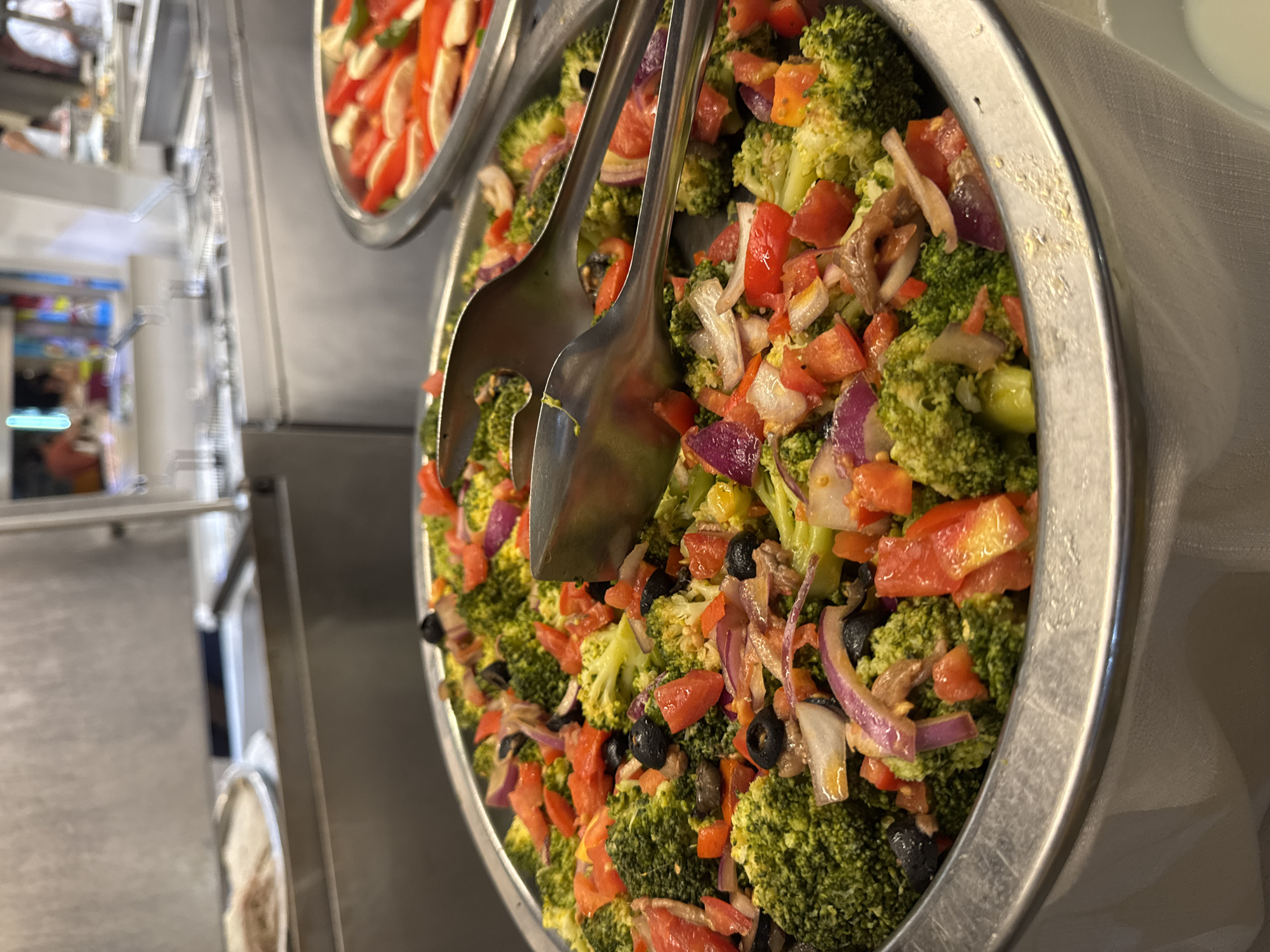 This is a close-up, eye-level, outdoor shot of a buffet food display, with a focus on a large metal bowl filled with a broccoli salad. The salad contains visible florets of broccoli, chopped red tomatoes, sliced black olives, and thinly sliced red onions. Two large, silver salad tongs are resting on top of the salad, with one partially submerged. In the background, another metal bowl containing what appears to be sliced red bell peppers and possibly other vegetables is visible. Behind that, a stainless steel buffet station with multiple compartments and a raised glass sneeze guard is present. The overall setting suggests a restaurant or catering establishment, likely in a warm climate. The city of Isola delle Femmine, Italy, is not depicted or implied in this image.
