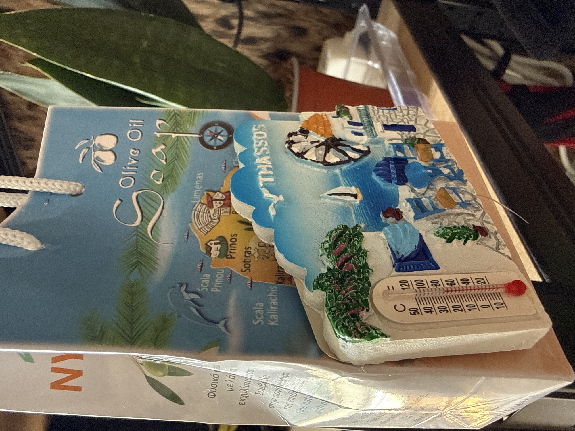 The image shows a souvenir set on a table or shelf, comprising two main elements. The first is a blue-toned cardboard package labeled "Olive Oil Soap," featuring a map of the island of Thassos, Greece, along with illustrations of olives, a compass, pine trees, and a dolphin. Various locations on the island are marked on the map. The second element is a 3D souvenir depicting a scene from Thassos, including a windmill, traditional buildings, and a thermometer showing a high temperature. The souvenir is in a plastic wrapping. The background is slightly out of focus, showing what looks like leaves and parts of other objects. There is no indication of the location being in Sofia, Bulgaria.