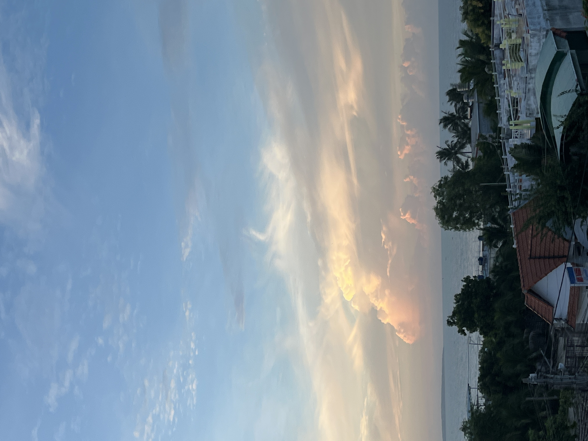 The image depicts a sky filled with soft, wispy clouds, transitioning from a clear blue on the left to hues of orange and pink on the right, suggesting either sunrise or sunset. Below the sky, a tranquil body of water, likely the sea, stretches towards the horizon. The foreground features a tropical landscape with lush green trees, including palm trees, and hints of buildings and structures. These structures appear to be part of a coastal settlement, with visible rooftops and some outdoor seating or balcony areas.
The overall scene evokes a sense of peace and natural beauty. The time of day is clearly indicated by the warm, diffused light illuminating the clouds, creating a serene atmosphere. While no people are explicitly visible, the presence of buildings suggests human habitation in the area. The location cues, such as the palm trees and the coastal setting, are consistent with a tropical environment like Mũi Né, Vietnam. There are no discernible signs with visible text.