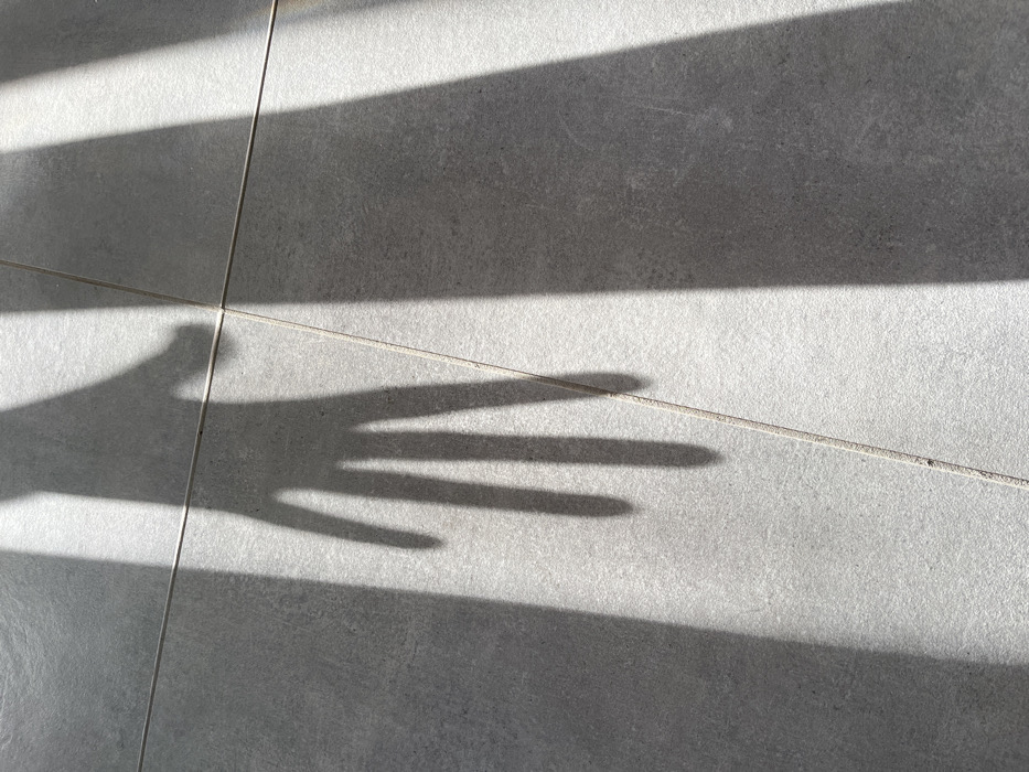 The image displays the shadow of an open human hand cast upon a light grey tiled floor in Chambœuf, France. The shadow, originating from an unseen subject, exhibits five distinct digits, with the thumb curled inward and the other four fingers partially spread. The flooring consists of rectangular, light grey tiles with visible, lighter-colored grout lines forming a grid pattern. Strong, directional light, likely sunlight, creates sharp, dark shadows, including the hand, and linear bands of shadow extending vertically across the frame, indicating light entering through a structured opening. No people or additional objects are directly visible within the illuminated areas or shadows.