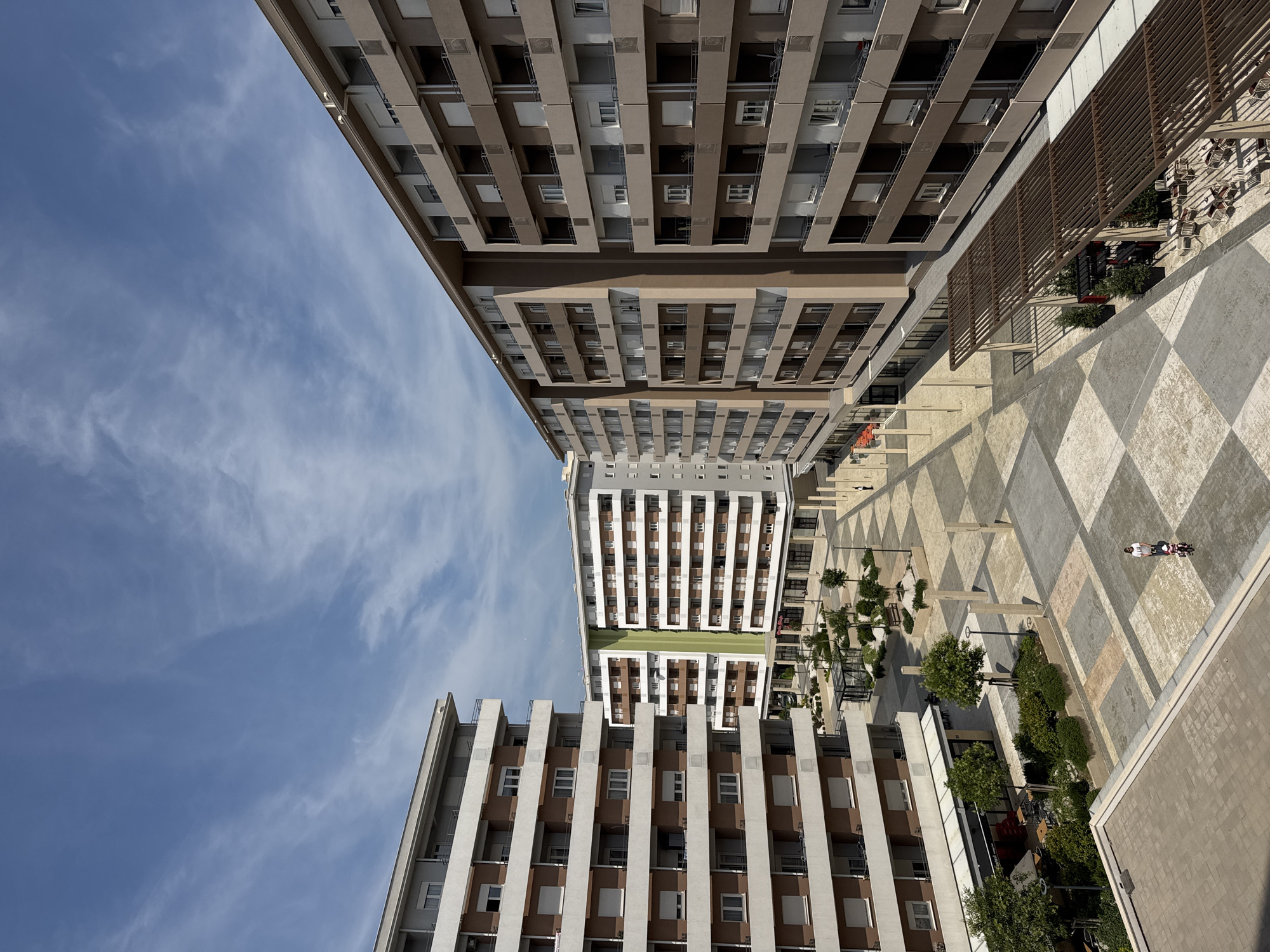 In an outdoor urban setting, a view looks down on a checkered paved plaza with several multi-story residential buildings surrounding it. The buildings, which vary in facade color and design, feature numerous balconies with railings and closed shutters on the windows. The sky above is a light blue with scattered white clouds. On the plaza, there are a few sparse trees, benches, and covered seating areas with tables and chairs, indicating a commercial or recreational space. A person is seen walking with a stroller on the plaza. The ground pavement is a mix of light and dark gray squares, with some areas of tan and off-white accents. Sunlight casts shadows across the scene.