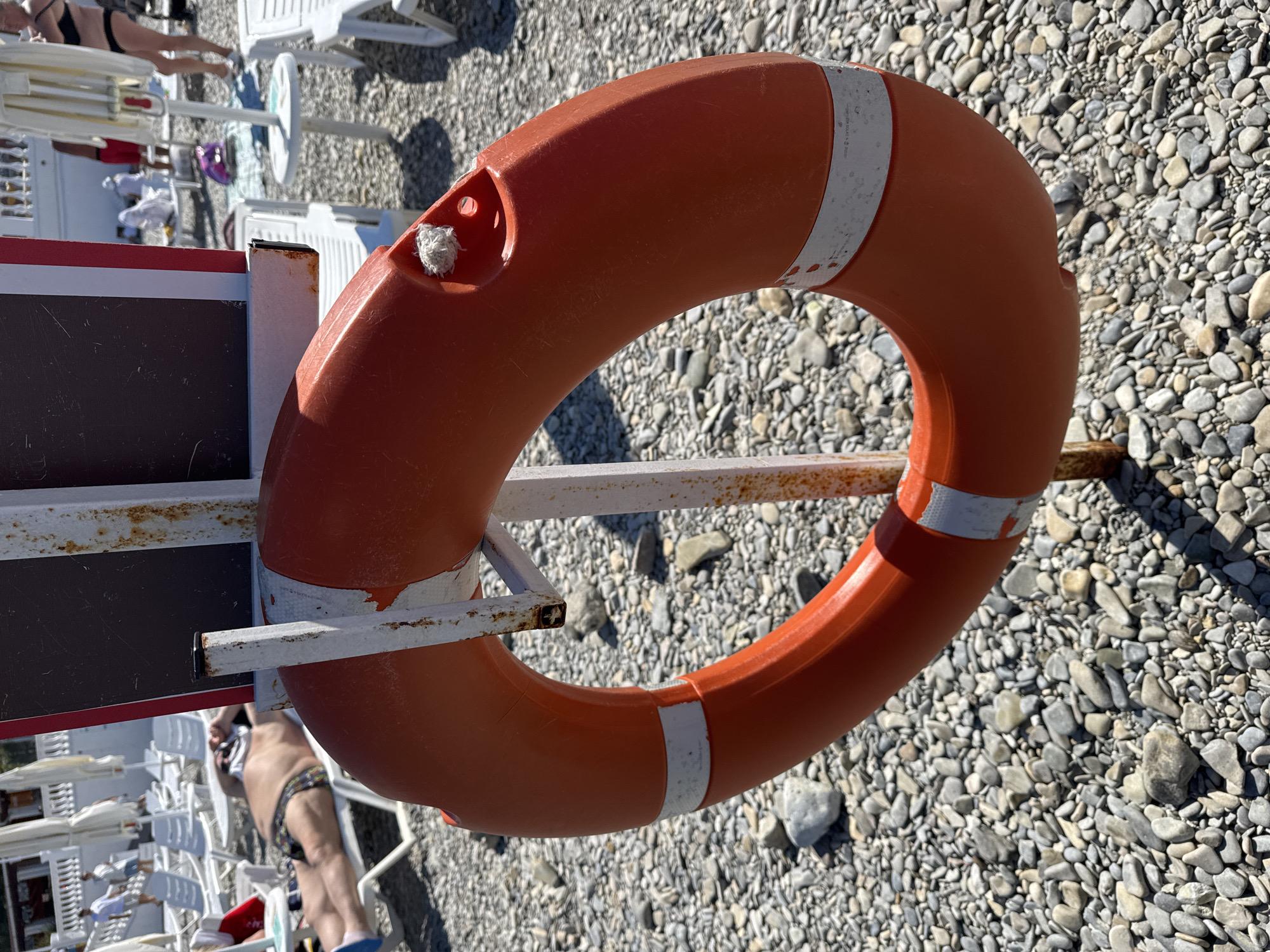 An orange life preserver hangs from a white metal pole on a pebble beach in Gelendzhik, Russia. The life preserver has two white reflective bands and a small piece of white rope sticking out of a hole. Behind the life preserver, lounge chairs, beach umbrellas, and several people are visible. The water is not visible in the frame.