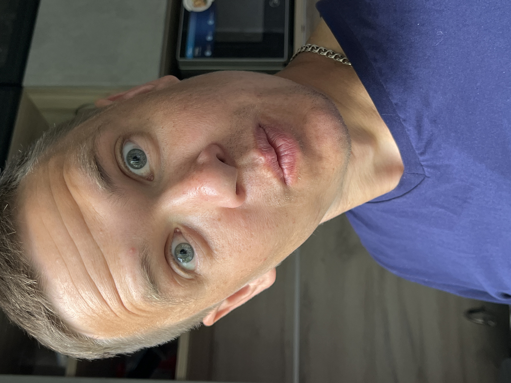 A close-up shot of a man with fair skin and short, light-colored hair. He has light blue eyes, visible wrinkles on his forehead, and a small amount of stubble on his chin. He is wearing a blue t-shirt and a silver chain. The background includes a gray microwave and a wooden surface. The location cannot be determined.
