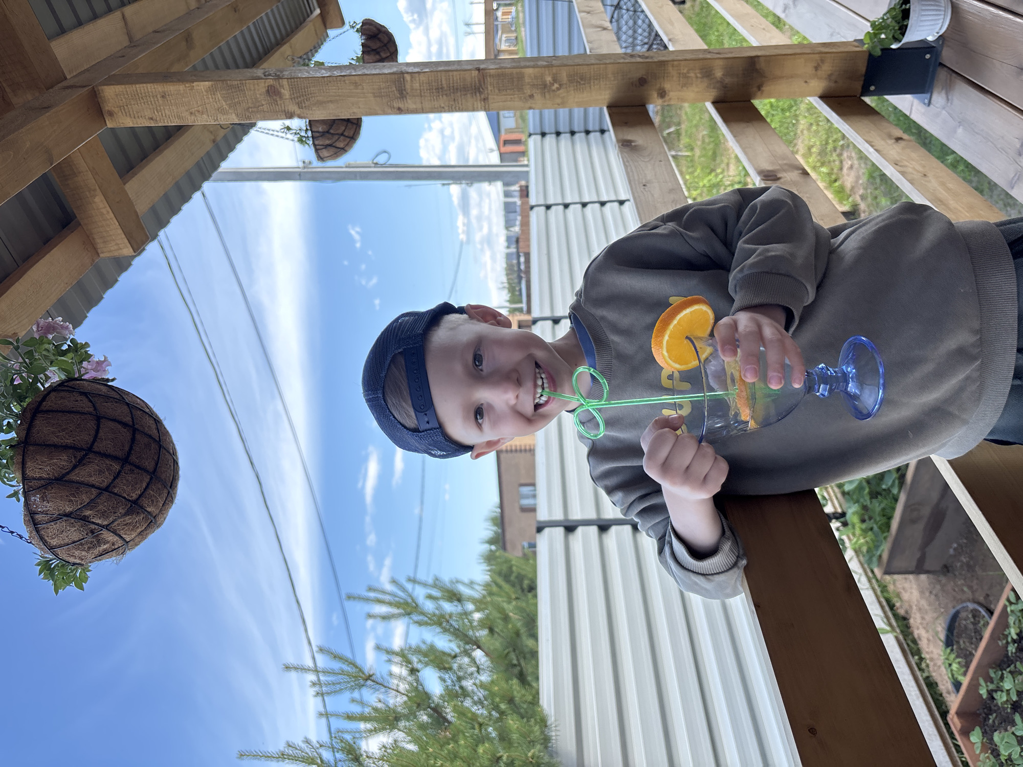 A young boy with light hair, wearing a dark blue baseball cap backward and an olive-green sweatshirt, stands on a wooden porch or deck in Chaikovsky, Russia, on a clear day with a blue sky and scattered white clouds. He is smiling widely, revealing a missing front tooth, and holds a blue stemmed glass containing a clear beverage with orange slices and mint leaves, with a green curly straw in his mouth. His left elbow rests on a wooden railing. Behind him is a light-colored corrugated metal fence, beyond which are evergreen trees and distant low-rise residential buildings. Overhead, a brown coir hanging basket with green plants is visible, and parts of a corrugated metal roof with wooden support beams can be seen. To the left, below the railing, are visible raised garden beds with small plants, possibly strawberries. To the right, on the wooden deck floor, is a small white pot with a green plant. Power lines stretch across the mid-ground sky.