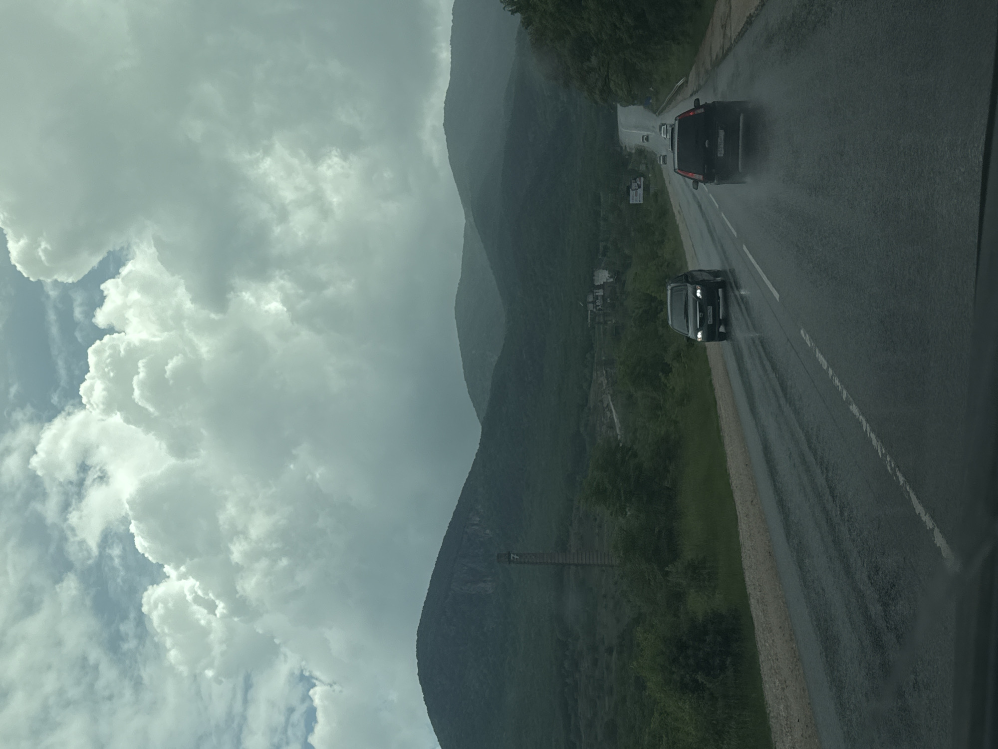The image depicts a road with two cars traveling on it. To the left, there's a view of mountains partially obscured by cloudy skies. The road is wet, suggesting it has been raining. Vegetation is visible on the side of the road between the road and the mountains. A structure resembling a bridge or walkway can be seen on the side of one of the mountains. The location is Unknown, Unknown.