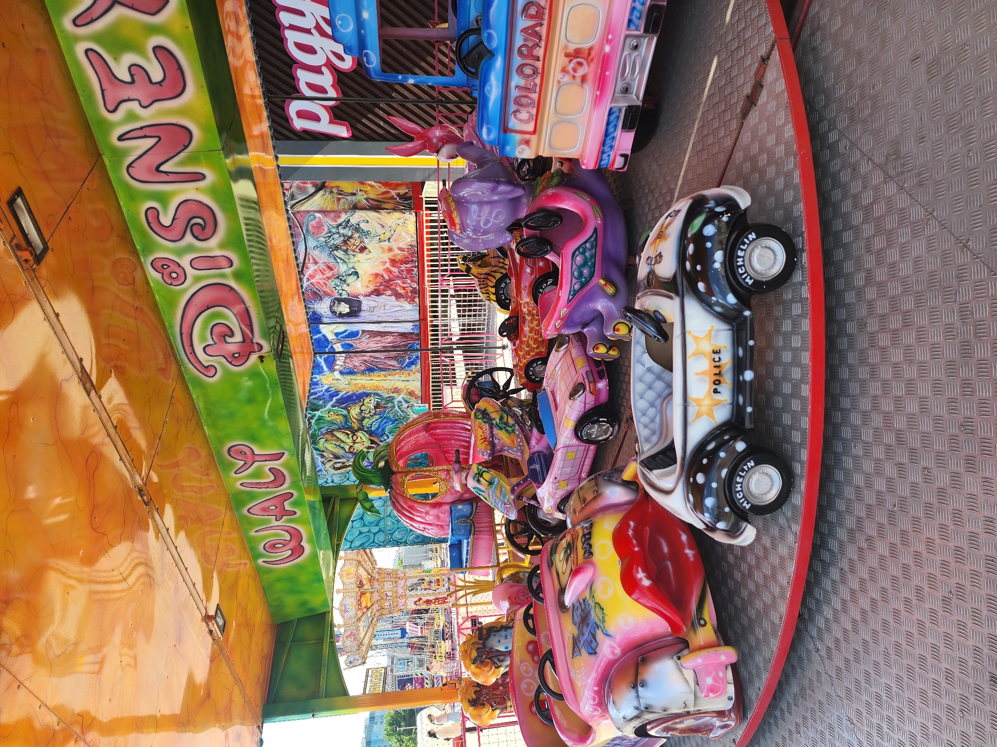 The image depicts a children's amusement ride featuring colorful, cartoonish cars and vehicles arranged in a circular formation on a metal platform with a red safety border. The cars include a police car, a Colorado-themed bus, and several other fanciful designs. Painted backdrops with vibrant illustrations surround the ride. Text on the ride reads "Waly Disney." In the background, glimpses of other carnival attractions and urban buildings suggest an outdoor amusement park setting. The location is Kazan, Russia.