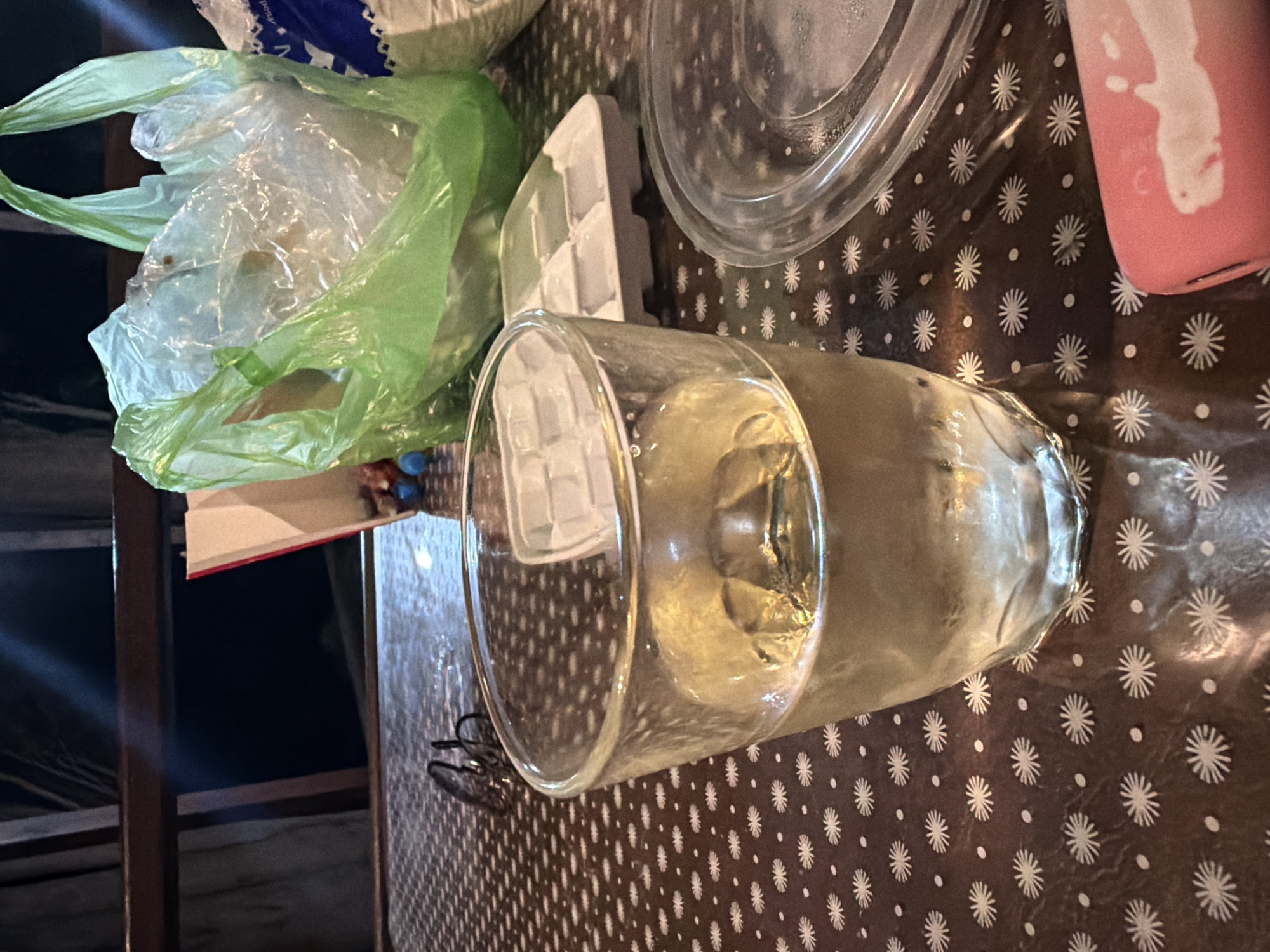 The image shows a close-up of a table with a glass of pale yellow liquid, likely a beverage, filled to about two-thirds of its capacity. The glass is made of clear glass and has a textured base. Next to the glass, an empty ice cube tray is visible. In the background, a green plastic bag, a book with a red cover, and a clear plastic lid or dish are present. The table is covered with a dark brown tablecloth featuring a pattern of white starbursts and small dots. A pair of eyeglasses rests on the tablecloth to the left of the glass. A portion of a pink object, possibly a phone or power bank, is visible in the lower right corner. The scene appears to be set outdoors, possibly on a balcony or patio, with dim lighting suggesting it is nighttime or indoors with low light. There is no indication of the city of Sochi, Russia, or any specific location.