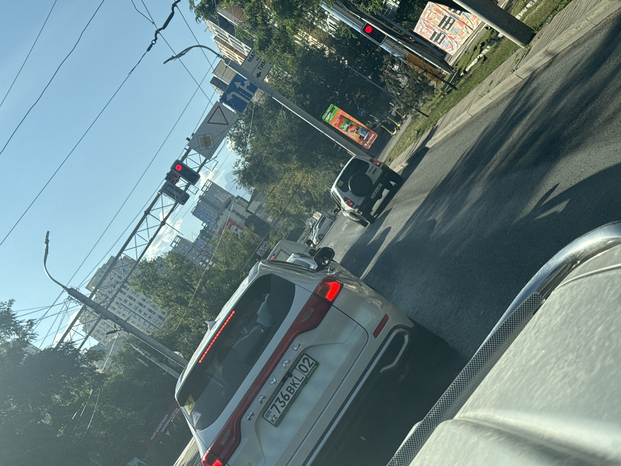The image shows a street scene in Almaty, Kazakhstan, likely on a sunny day. In the foreground, the hood and windshield of a car are visible from the driver's perspective. Following the road, we see the rear of a silver Haval SUV with a license plate reading "KZ 736 BKL 02". Further down the road, other vehicles are present, including a white SUV with a spare tire mounted on the back. The scene is characterized by urban infrastructure, including traffic lights, street signs, and power lines overhead. Tall buildings are visible in the background, suggesting a developed urban area. Trees line the sidewalk, and there is some greenery present. An advertisement for "Juice" can be seen on a billboard along the roadside. The overall impression is of a typical city street with moderate traffic.