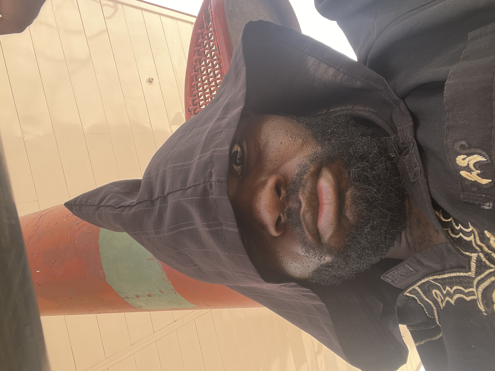 A close-up, low-angle, rotated view depicts a Black male wearing a dark, hooded jacket. The hood partially obscures his face, revealing one eye, his nose, and a full beard. His jacket appears to have embroidery on the chest area. In the background, there's a curved pole painted with alternating red and green sections, and a white wall with vertical paneling. The location in Barakin Lerawi, Nigeria cannot be determined from this image.