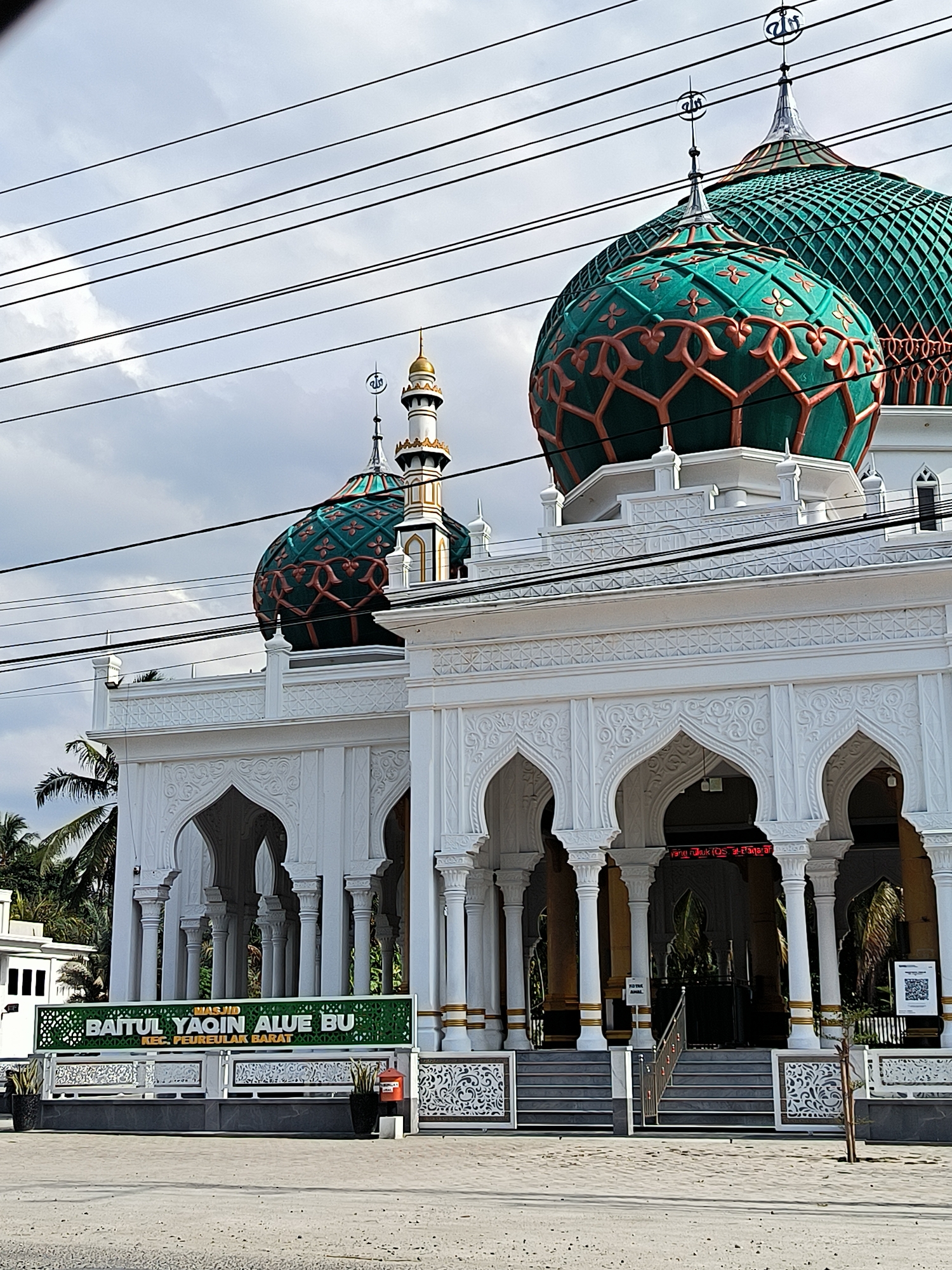 The image shows the Baitul Yaqin Alue Bu mosque in Aluebutuha, Indonesia, in the Peureulak Barat district. The mosque is a white building with arched entrances and columns, topped with distinctive green and orange-patterned domes. A minaret is visible beside one of the domes. A green sign in front of the building displays the mosque's name in English: "BAITUL YAQIN ALUE BU, KEC. PEUREULAK BARAT". A set of stairs leads up to the mosque entrance. Power lines are visible against the cloudy sky in the background.