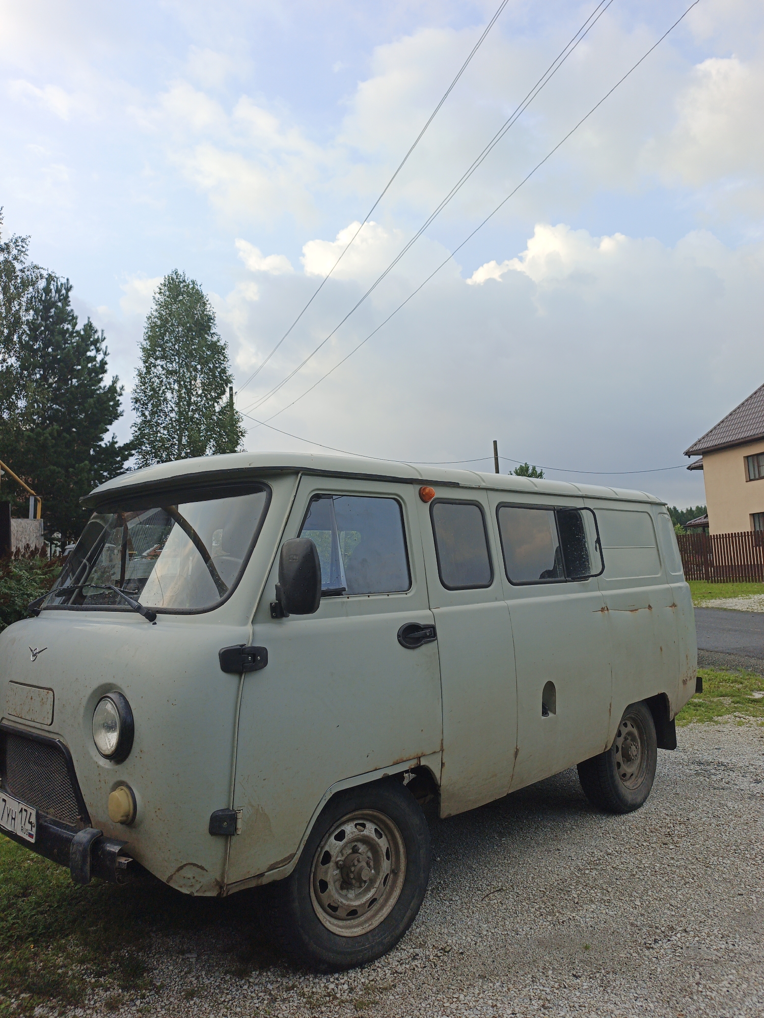 This image shows a light green UAZ-452 van parked on a gravel driveway in what appears to be a rural or semi-rural setting. The van is the primary subject, occupying the center of the frame. It is an older model, showing signs of wear and tear, including rust spots on its exterior. The van has a boxy shape with multiple windows along its side and a visible orange indicator light on the roof.
Behind the van, there is a residential building with a brown tiled roof and a wooden fence. Trees, including coniferous and deciduous varieties, are visible to the left and in the background. Power lines are strung across the sky, suggesting the presence of infrastructure.
The sky is partly cloudy, with a mix of blue sky and white clouds, indicating it is likely daytime, possibly late afternoon or early evening given the lighting. The ground is covered in gravel and some patches of grass. The overall impression is that of a quiet, ordinary day in a village or small town. There is no visible activity or event taking place; the van is simply parked.
The license plate on the front of the van is partially visible and appears to read "7 YH 174 RUS". The emblem of UAZ is visible on the front of the van. The location context provided is Medvedyovka, Russia.