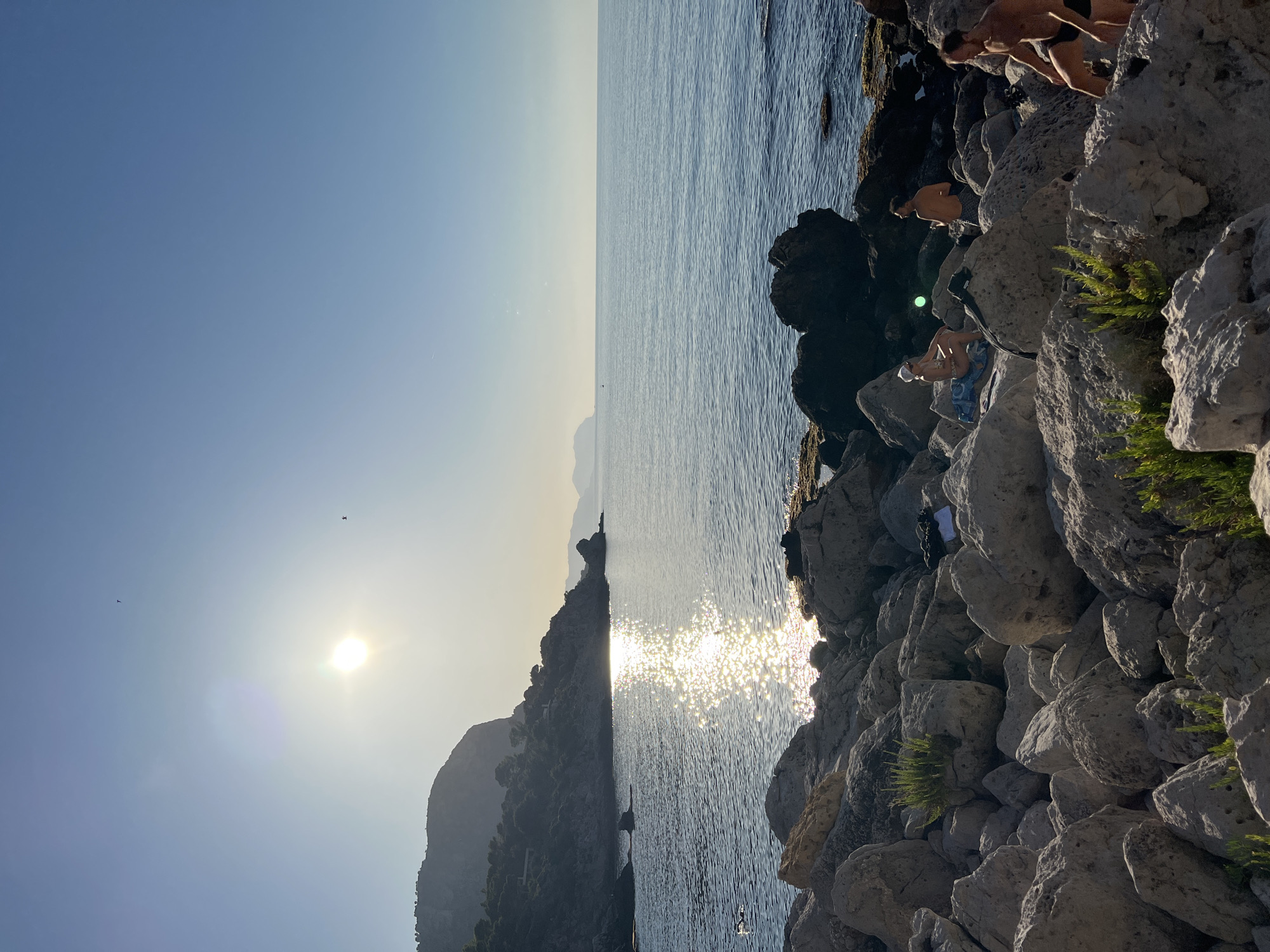 The image shows a rocky coastline with the sea on a clear day. The sun is setting or rising, casting a strong reflection on the water. Several people are present on the rocks. In the foreground, three men are visible. One man is partially visible on the right side of the frame, walking and wearing dark swim trunks. Another man stands slightly to the left, also wearing dark swim trunks. Further left, a woman with a white hat is sitting on a towel, facing the sea. The coastline is characterized by large boulders and some sparse vegetation. In the background, a hillside covered in trees and some structures rises from the sea, with the silhouette of a mountain range on the horizon. The sky is clear blue with a soft haze from the sun.