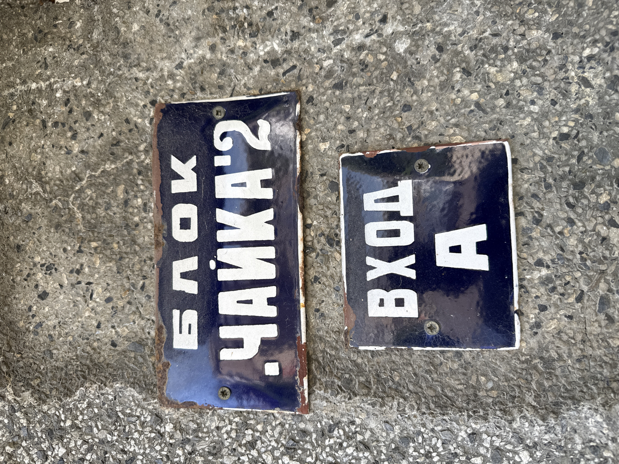 The image displays two weathered, rectangular enamel signs, dark blue with white Cyrillic text, securely fastened to a coarse-grained concrete surface in Kardzhali, Bulgaria. The upper sign, positioned above the lower one, reads "БЛОК .ЧАЙКА'2" (BLOCK .CHAIKA'2, or BLOCK .SEAGULL'2). It exhibits considerable rust along its perimeter, particularly on the top and bottom edges, and minor chipping of the enamel surface. Two screws are visible in its upper left and lower right corners, securing it to the concrete. The lower sign, smaller in size, states "ВХОД А" (ENTRANCE A) and also shows rust and enamel deterioration, predominantly along its bottom edge. Two screws are visible in its upper corners. The background concrete surface is light gray, textured with visible aggregate, suggesting a wall or a paved outdoor area. No people or actions are depicted in the scene.