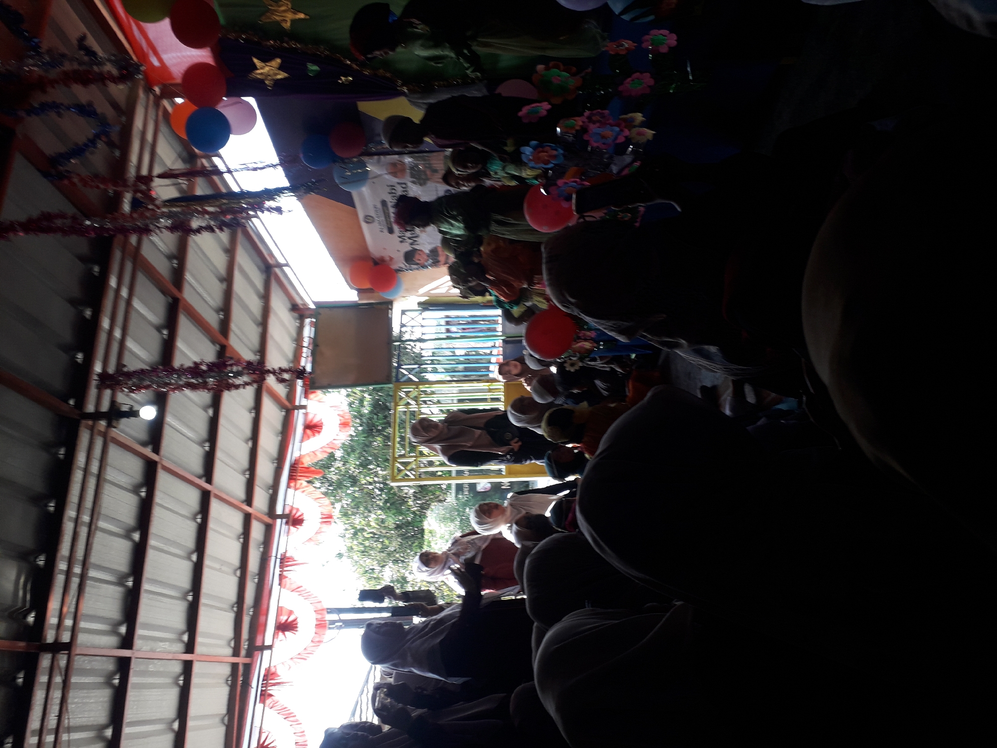 The image depicts an outdoor gathering under a covered structure with a corrugated metal roof, likely in Cankuang, Indonesia. A group of individuals, predominantly women and children, are present. Many of the attendees are wearing hijabs, suggesting a cultural or religious context. Some individuals are standing and appear to be observing or participating in an event, while others are seated or partially visible in the foreground, with their backs to the camera, creating a silhouette. In the background, there's a yellow metal gate and some foliage, hinting at an open or semi-open space. Decorations such as balloons, tinsel, and star-shaped ornaments are visible, indicating a celebratory or festive occasion. A banner with Indonesian text is also present. The lighting suggests it is daytime, with sunlight filtering through the structure and illuminating parts of the scene.