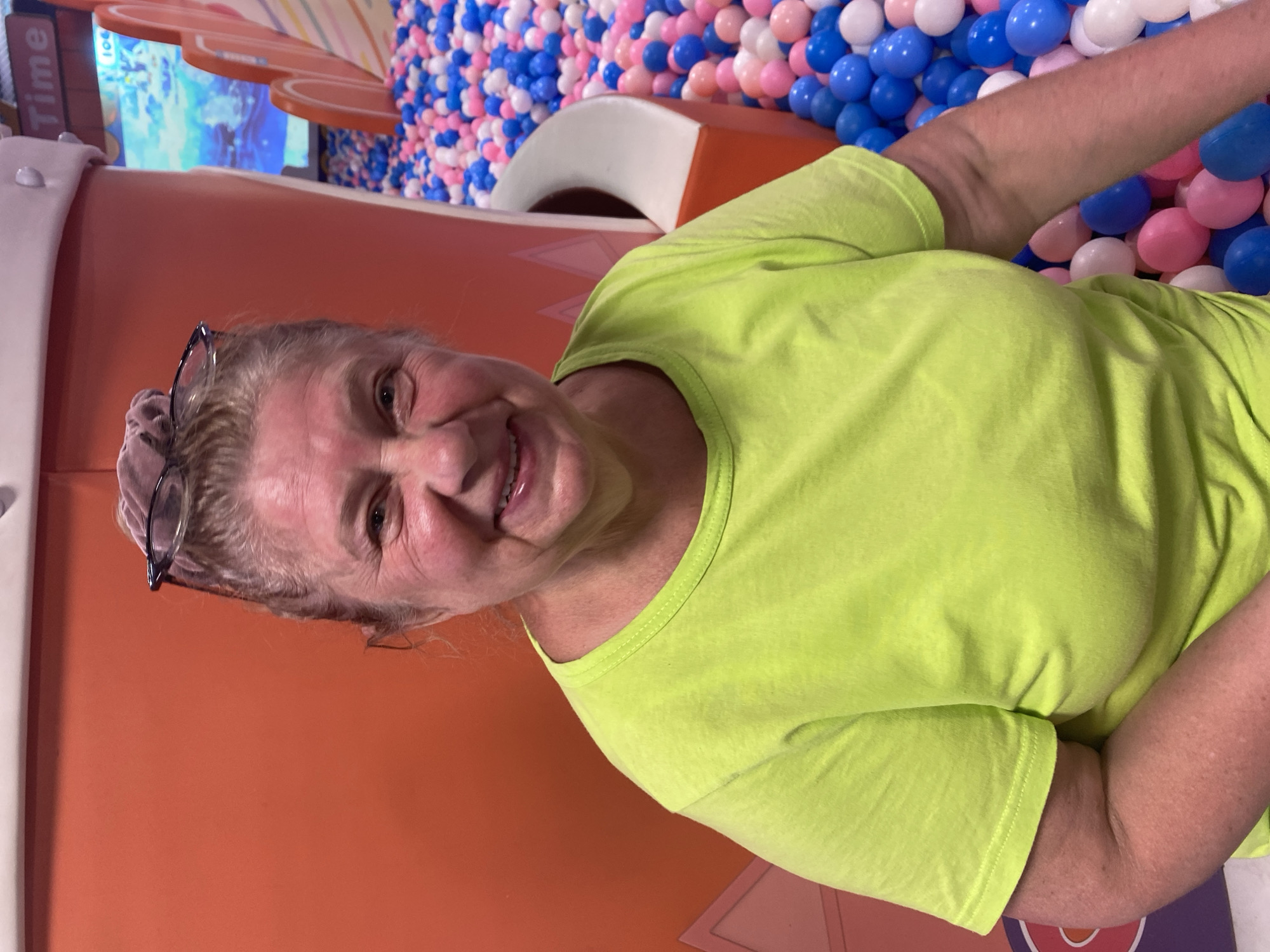 An older woman with blonde hair pulled up and glasses perched on her head smiles at the camera. She is wearing a bright green t-shirt and is seated in front of an orange wall. To her right, a ball pit filled with pink, blue, and white balls is visible. A screen displaying a video game or animation is also visible in the background, with the word "Time" displayed at the top. The environment suggests an indoor entertainment venue, possibly a play area or arcade. The woman's location in Phu Thuy, Vietnam is not indicated by any visual cues in the image.