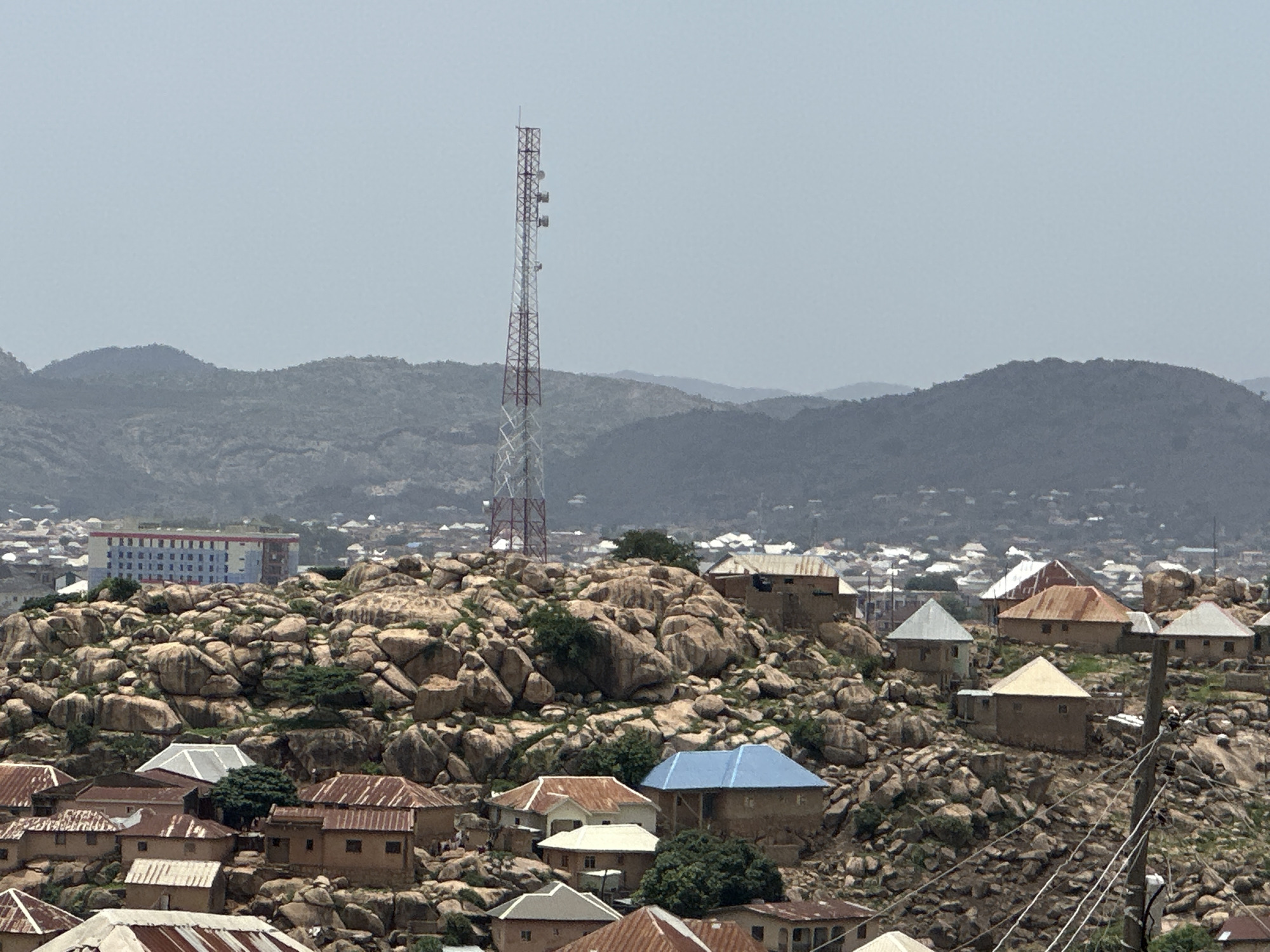 The image shows a view of a neighborhood in Jos, Nigeria, characterized by rocky terrain and traditional houses. A red and white telecommunications tower stands prominently on a rocky outcrop overlooking the community. The houses are a mix of styles, some with rusted corrugated iron roofs, while others have blue or white roofs. The houses are interspersed with large boulders and sparse vegetation. In the background, a city landscape with more buildings and distant hills are visible under a hazy sky.