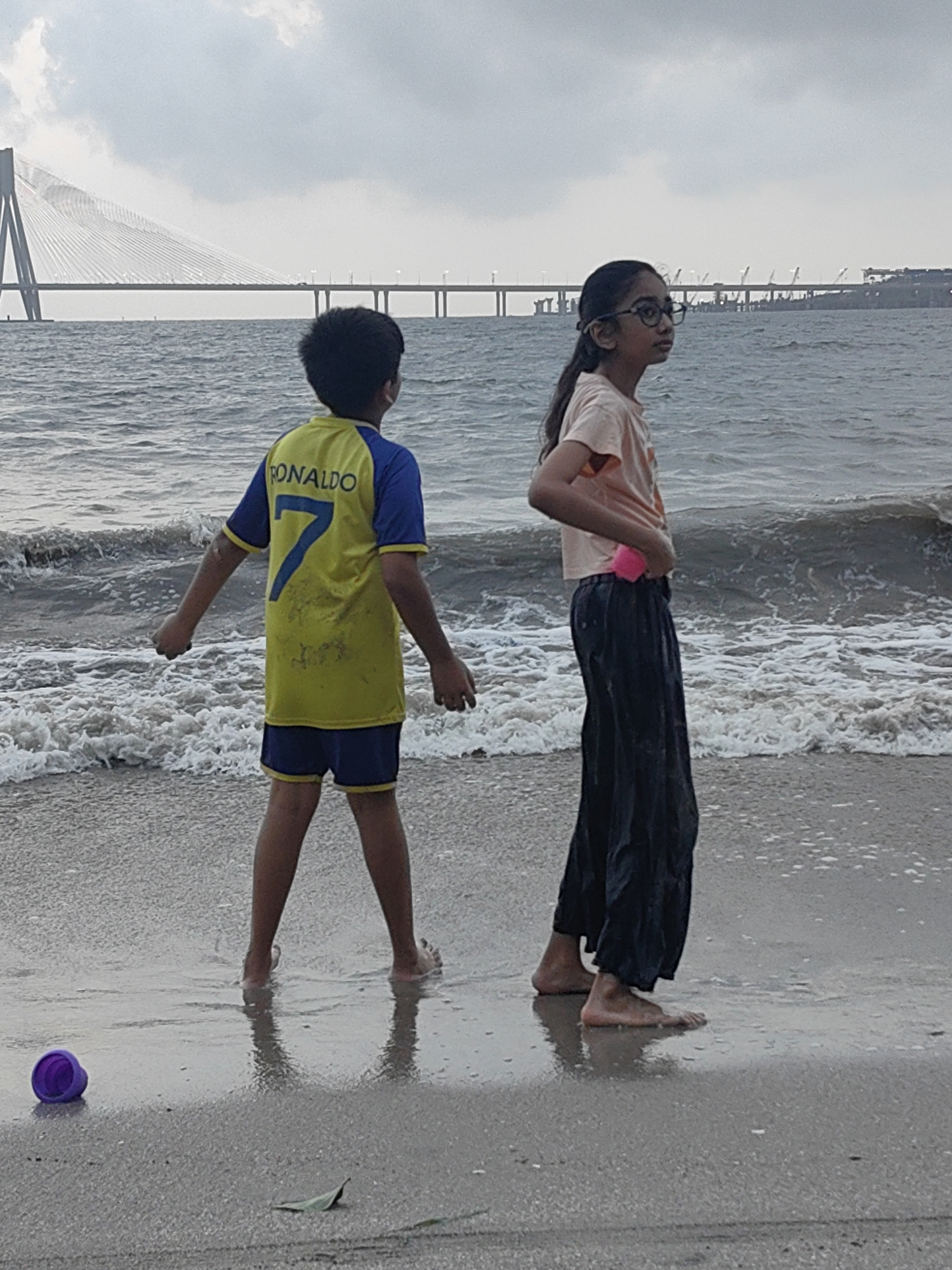 The image shows a boy and a girl standing on a beach in Mumbai, India, with a body of water and a bridge in the background. The boy, facing away from the camera, is wearing a yellow and blue soccer jersey with "RONALDO" and the number "7" printed on the back, and blue shorts. The girl, facing to the side, is wearing a light orange t-shirt and dark pants. They are both standing near the water's edge, with waves lapping at their feet. A purple toy bucket lies on the wet sand to the left of the boy. The sky is overcast, and the overall lighting suggests it is daytime. In the distance, a long, modern bridge spans across the water, characteristic of Mumbai's infrastructure, possibly the Bandra-Worli Sea Link. The environment is a typical urban beach setting.
