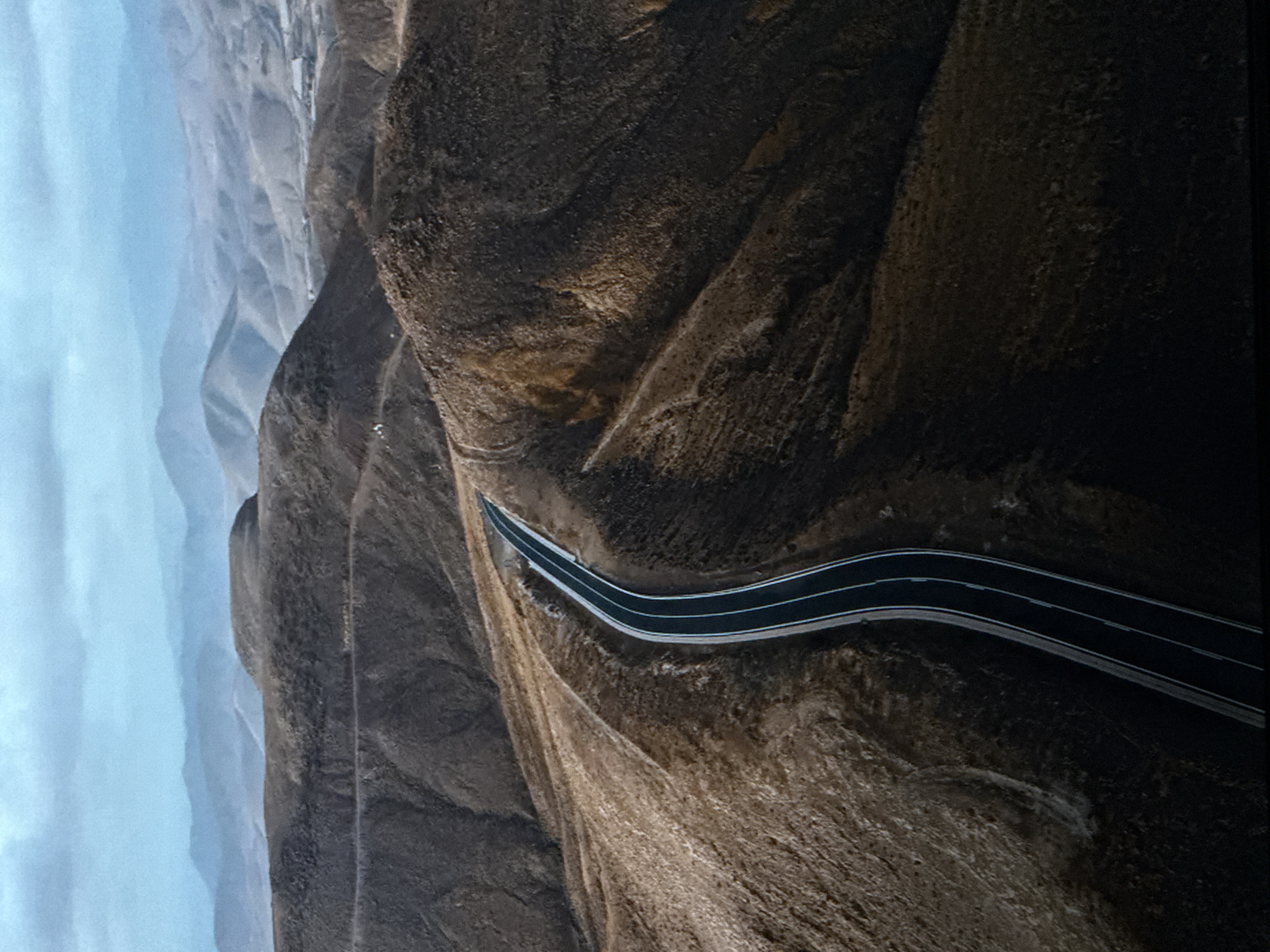 The aerial photograph captures a winding asphalt road traversing a desolate, mountainous terrain. The road, marked with white lines, curves through the rugged landscape, with steep, barren hillsides on either side. The hills are covered in dry, brown vegetation and show signs of erosion. In the distance, layers of hazy mountains recede into the horizon under a cloudy, pale blue sky. The overall impression is one of a remote and rugged environment. There are no people, animals, or specific landmarks visible that would associate this image with the city of Saratov, Russia.