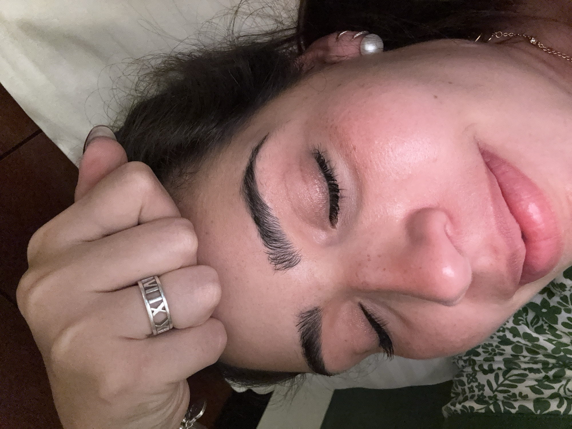 A close-up, slightly angled shot depicts a person lying down with their eyes closed. They have well-defined eyebrows and eyelashes. Their hand is resting on their face, and a silver ring with Roman numerals is visible on their finger. The background consists of a patterned green and white fabric. The location is indeterminate.