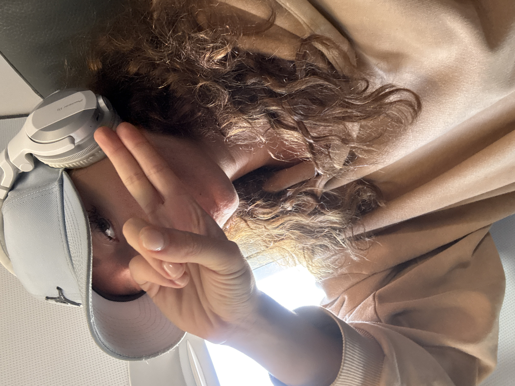 A person with curly brown hair, wearing a light grey baseball cap with a dark logo on the front and white headphones around their neck, holds up three fingers in front of their face. The person is wearing a tan-colored hooded sweatshirt. The background shows parts of an airplane interior, including a window and wall. The location of the image is not discernible as being in Nong Prue Subdistrict, Thailand, or any specific city.