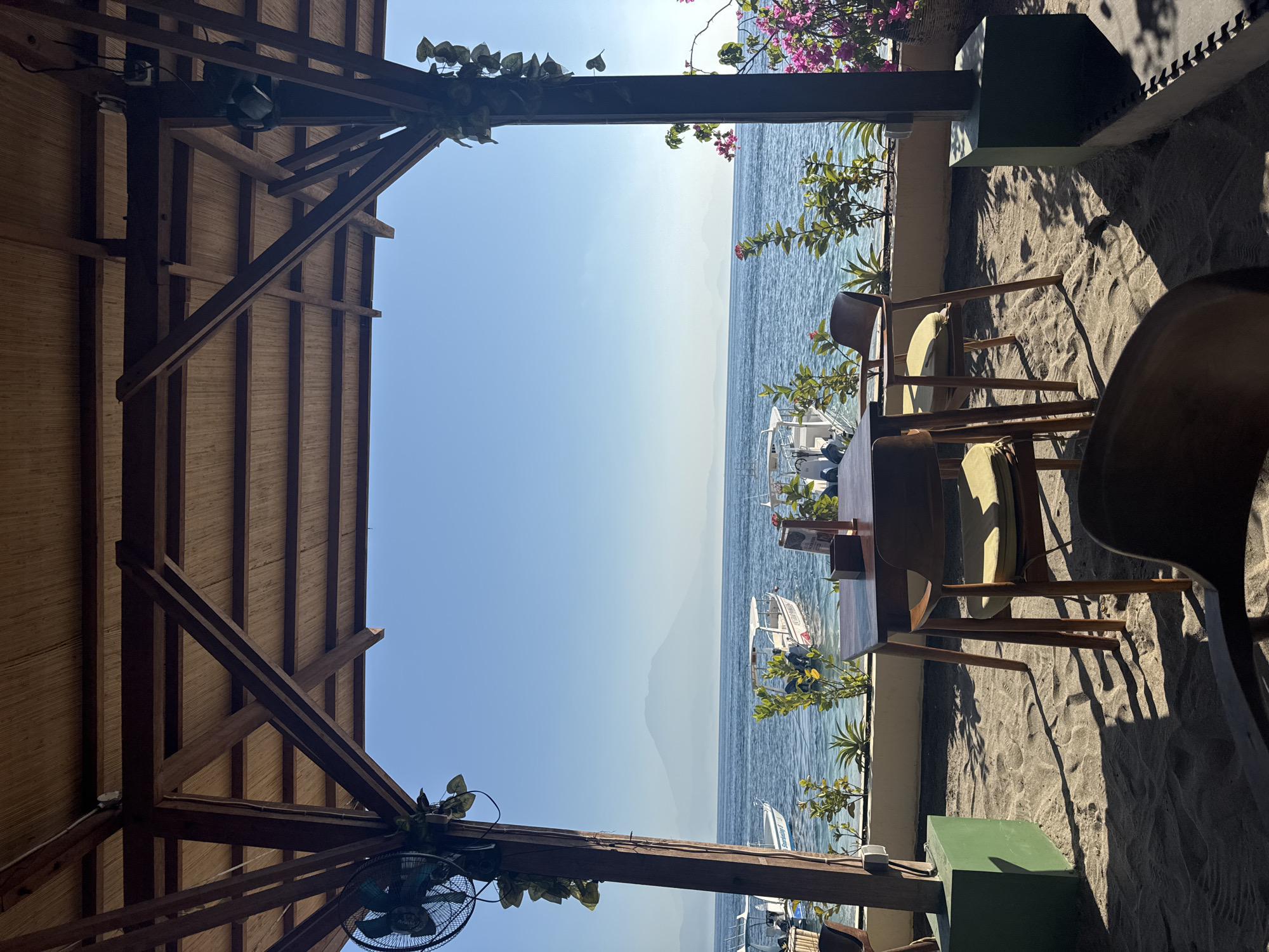 The image displays an open-air, beachside establishment, likely a restaurant or cafe, situated on Nusa Penida, Indonesia, providing a direct view of the ocean and the distant Mount Agung on Bali. The scene is brightly lit by natural sunlight, casting sharp, elongated shadows across the sandy floor, indicating a clear day.
The foreground is composed of a light brown sandy surface, showing discernible footprints and the shadows of furniture and the overhead structure. A rectangular dark wooden table with two chairs, each featuring light green cushions, occupies the mid-ground. A portion of a third, darker wooden chair is visible in the immediate foreground, its back facing the viewer. On the table, small wooden containers, possibly holding napkins or condiments, are present, alongside what appears to be a menu or informational sign.
The overhead structure of the establishment consists of dark brown wooden beams and supports, beneath a ceiling made of a lighter, woven natural material, possibly bamboo matting. Exposed electrical wiring and a small white fixture are visible on the ceiling. Two substantial vertical wooden pillars anchor the structure, framing the expansive view. Attached to the left pillar is a dark green caged fan, partially obscured by green trailing vines. Similar green vines cascade from the right pillar, which is accompanied by a vibrant plant displaying numerous pink bougainvillea flowers. A dark woven basket is partially visible behind the pink flowers. Both pillars terminate in dark green, cube-shaped bases resting on the sand.
Beyond the shaded area, the calm, blue ocean extends towards the horizon. Several small white motorboats are scattered across the water, appearing to be anchored or docked near the shore. One boat on the left clearly displays "Seruling 5" on its side, while another on the right has some red lettering. A narrow strip of low-lying green plants and small shrubs with red flowers forms the immediate shoreline, separating the sandy patio from the water.
In the far distance, a large, hazy, conical mountain, identifiable as Mount Agung on Bali, dominates the central horizon against a clear, light blue sky. To its left and right, fainter, more distant landmasses are also visible through the atmospheric haze.