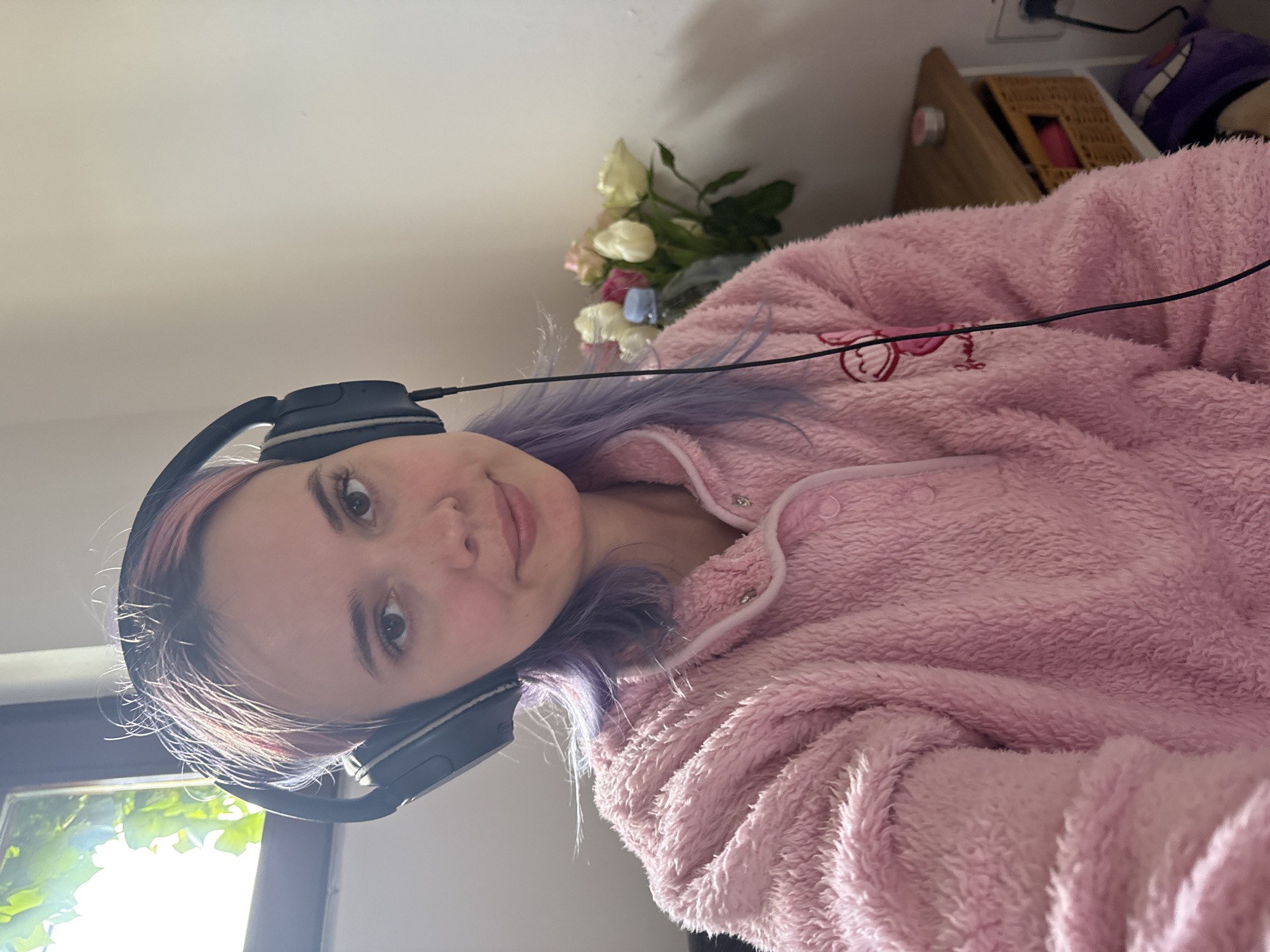 A young adult female is depicted indoors in the City of Zagreb, Croatia. She has light skin, brown eyes, and hair dyed pink and purple, with visible lighter roots. She is wearing dark over-ear headphones and a pink, fuzzy long-sleeved pajama top with white trim, snap button closures, and a subtle embroidered character emblem on the chest. She faces the viewer with a slight smile. A window, showing bright light and green foliage, is behind her to the left. To her right, a bouquet of white, pink, and yellow roses is arranged in a vase on a light-colored wooden corner table. The table also contains a wicker basket drawer and a small, light-colored cylindrical container. A white electrical outlet with a dark plug and a partially visible purple plush object are in the lower right foreground.