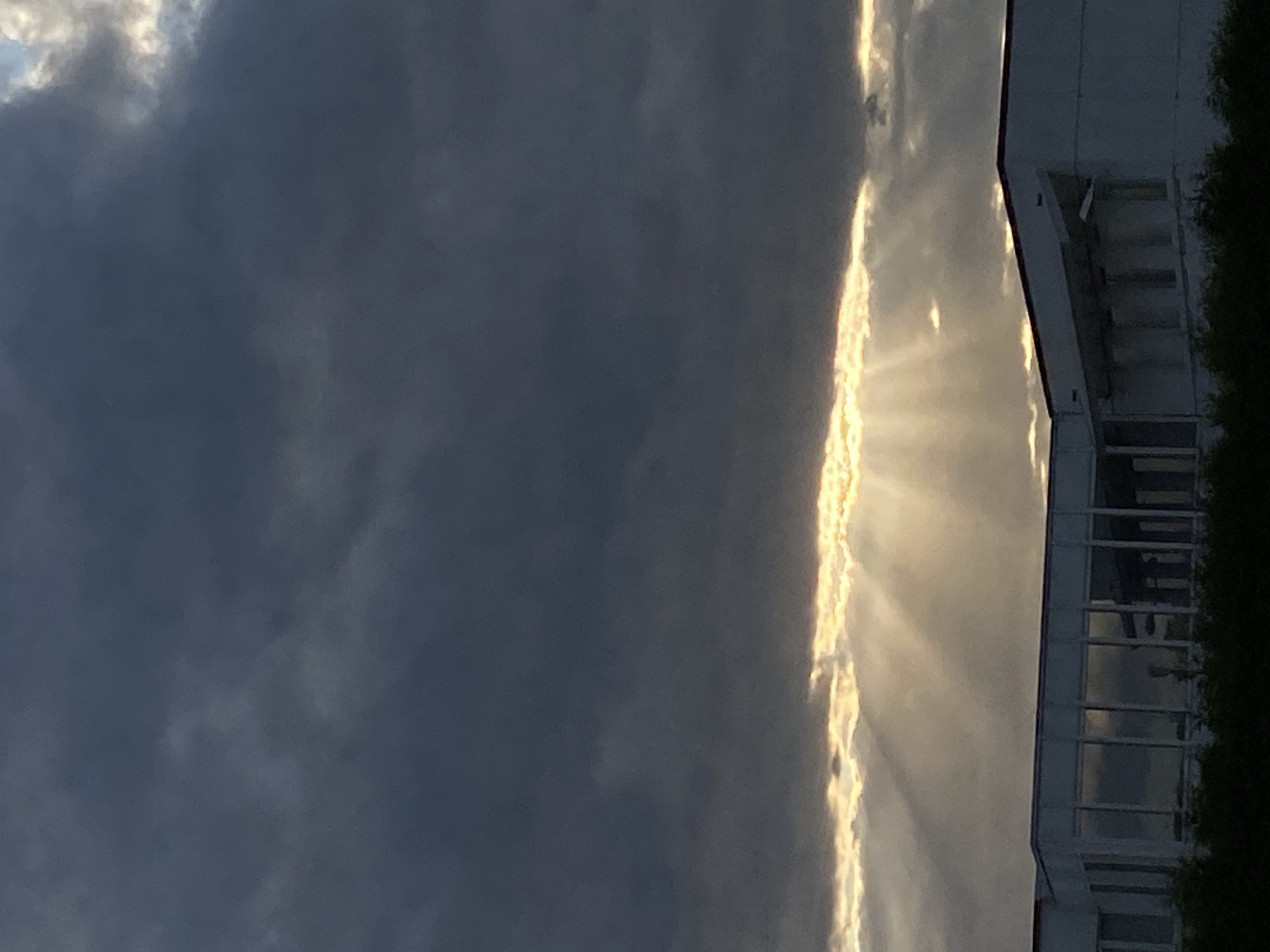 The image depicts a low-angle shot of a building's facade against a dramatic sky in Rinteln, Germany. The sky is filled with dark, heavy clouds, but a bright band of sunlight pierces through them, creating crepuscular rays that illuminate the scene. The building appears to be modern, with a white exterior and large, rectangular windows. Some of the windows reflect the stormy sky, adding to the dramatic atmosphere. A line of dark green foliage runs along the bottom of the image, partially obscuring the base of the building. No people or specific actions are visible in the image.