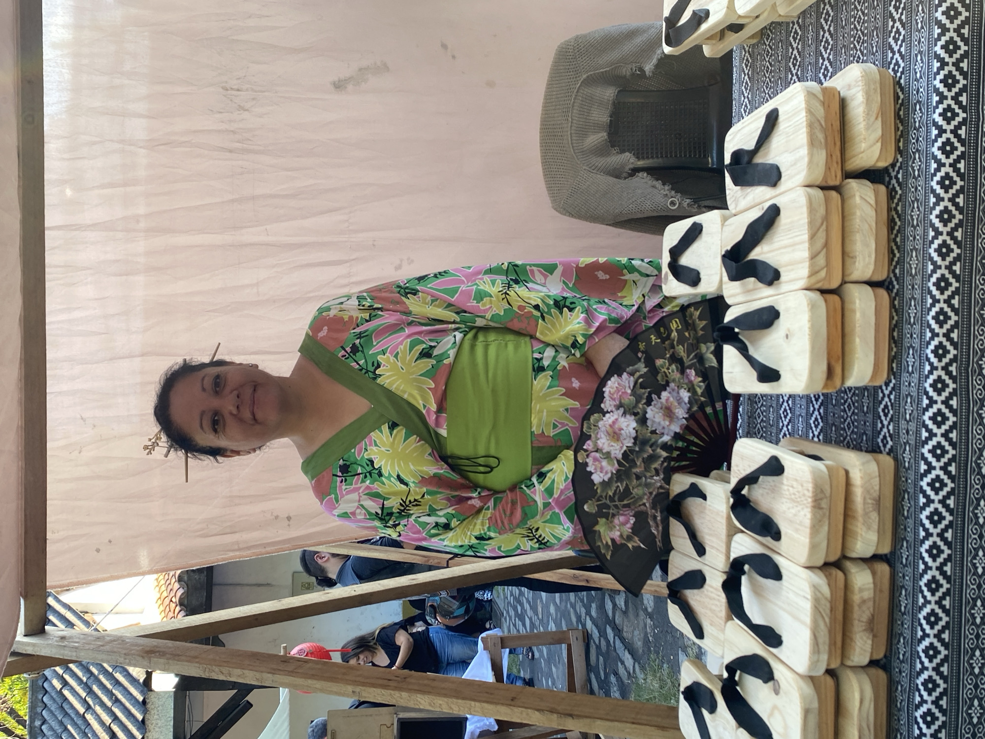 A vendor, a woman with dark hair and skin, wearing a colorful floral kimono, stands under a tent at a market stall in Buenos Aires, Argentina. She is holding a black fan with white and pink flowers. The stall displays numerous pairs of wooden geta sandals with black straps. The tablecloth is black and white with geometric patterns. In the background, blurred figures of other people and buildings can be seen. A wooden structure of the market stall frames the left side of the image.