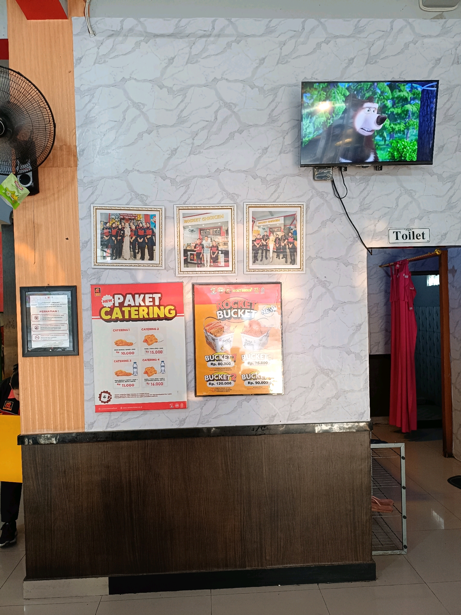 The image captures an indoor scene, likely a restaurant or shop, with a focus on a wall decorated with faux marble wallpaper. On this wall, a flat-screen television displays a cartoon image of a bear. Below the television, three framed photographs are hung in a horizontal row. The leftmost photograph shows a group of people in uniforms. The middle and rightmost photographs also depict groups of people, with the rightmost one appearing to be a staff photo.
Dominating the lower half of the wall space are two large posters. The left poster is red and white and advertises "PAKET CATERING" with various package options and prices. The right poster, also red and white, advertises "ROCKET BUCKET" meals, with images of fried chicken and prices. To the right of these posters, a doorway is partially visible, draped with a bright pink curtain, and a sign above the doorway reads "Toilet."
To the left of the posters, a black framed sign with white text is mounted on a wooden panel. A large black oscillating fan is mounted on the wooden panel to the left of the framed sign. The floor is tiled, and a dark brown wooden counter runs across the bottom of the image, with a metal shelving unit partially visible to the right.
The overall impression is a casual establishment that prioritizes visual information and customer service, with catering and meal deals prominently displayed. The presence of a television showing entertainment and a designated toilet area suggest it is a place for patrons to dine or relax. The location is identified as Ngemplak, Indonesia.