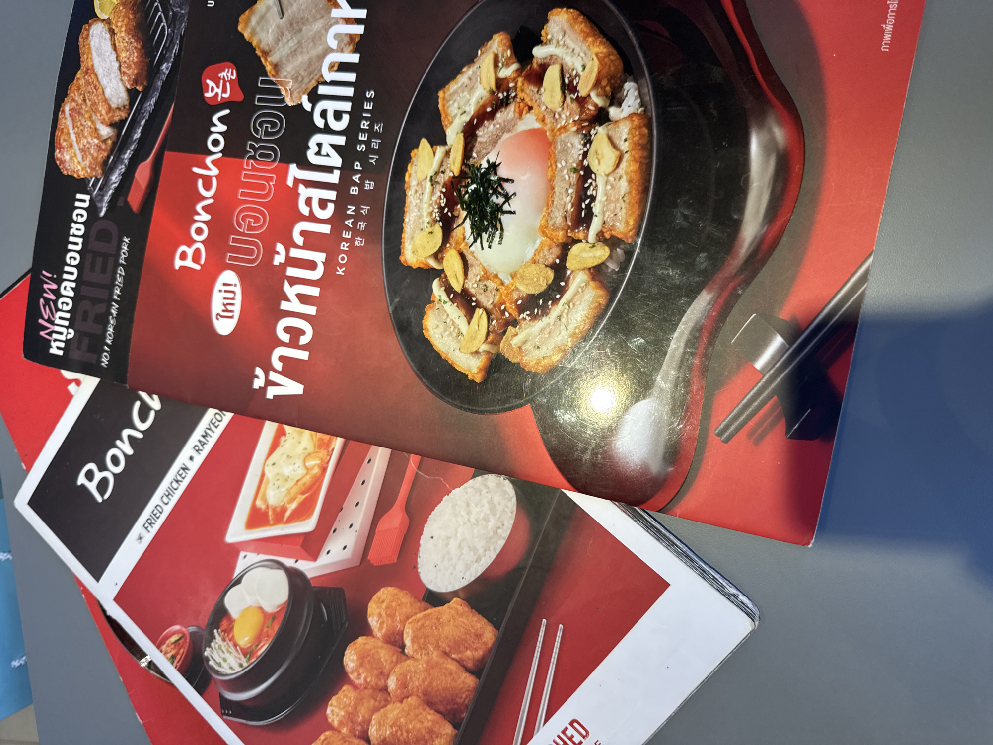 Two menus are laid out on a grey surface, overlapping each other. The menu on the left is mostly red with white text and images of food. It features the text "BonChon" and images of fried chicken and ramyeon. The menu on the right is red and black, with white and red text. It displays the text "Bonchon" and "NEW!" in Thai script, along with a large image of a dish with pork, rice, and a poached egg, topped with fried garlic slices and seaweed. The Thai text under "Bonchon" appears to be "본촌," and the text below the main dish image reads "KOREAN BAP SERIES" and then smaller Thai text followed by Korean text. The overall scene suggests a promotional display or casual dining setting, possibly in a restaurant in Bangkok, Thailand, given the Thai language present on one of the menus.