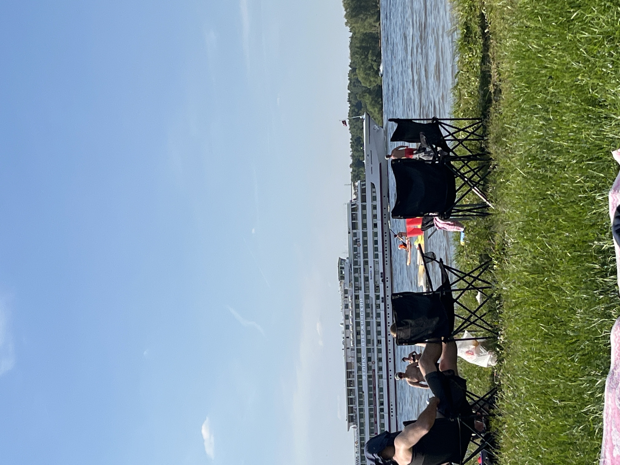 The image depicts a sunlit recreational scene along a body of water in Kapustino, Russia. In the immediate foreground, lush green grass occupies the bottom portion of the frame. On this grassy bank, a person is seated from the back in a black folding chair, wearing a dark top and shorts, with a blue fabric covering their head. To their right, another black folding chair is partially visible, occupied by an individual whose head is obscured. A white plastic bag rests on the grass between these chairs. Further right, a third black folding chair is seen, draped with a pink towel.
In the mid-ground, a large body of water, likely a river or lake, stretches across the frame. Several individuals are visible in the water near the shore; two men are standing, one shirtless in red shorts, and a woman in a dark swimsuit is partially submerged. Further out, a person is on a stand-up paddleboard, and a small yellow watercraft is near the right shore with an occupant wearing an orange vest. A small white watercraft is creating a wake in the far right mid-ground.
Dominating the mid-ground is a large, multi-decked white passenger ship, identified by the Cyrillic text "ФЕЛИКС ДЗЕРЖИНСКИЙ" (FELIX DZERZHINSKY) on its side. The ship features numerous windows and a red stripe along its hull. A dark red flag is flying from its bow as it moves from left to right.
The background consists of a clear, bright blue sky with sparse, wispy white clouds and a faint contrail. A dense line of green trees forms the distant bank of the waterway on the right side of the image. The overall scene suggests a warm, clear day with recreational activity along the waterfront.