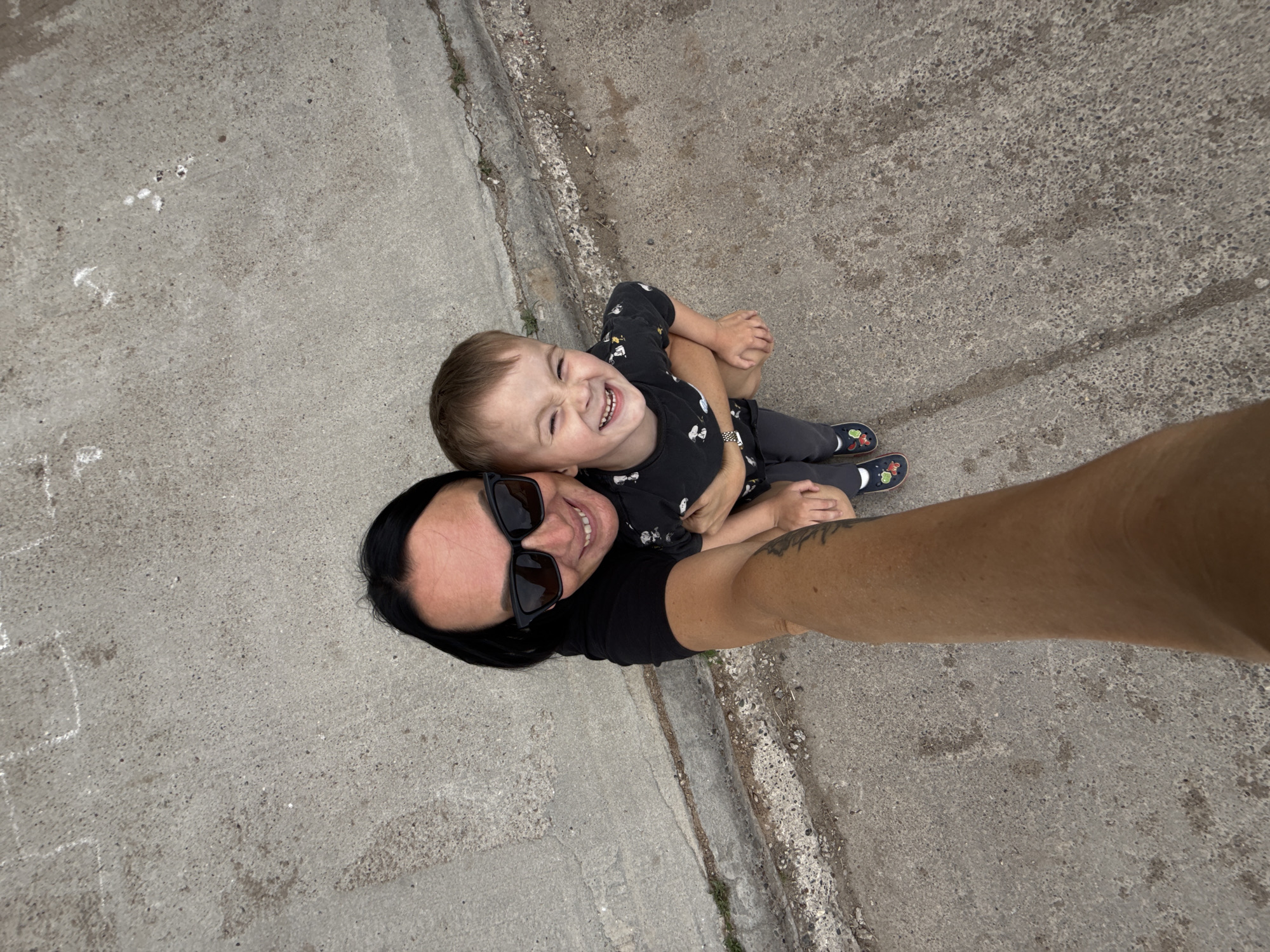 A woman wearing sunglasses and a black t-shirt is taking a selfie outdoors while holding a young boy. The boy is wearing a black t-shirt with a cartoon pattern and grey pants, and he is smiling widely. They are standing on a concrete surface, which appears to be a sidewalk or pathway. The background shows more of the concrete surface and some sparse vegetation along a crack. The overall setting suggests a casual outdoor environment, possibly a park or a street in Chaikovsky, Russia, given the context.