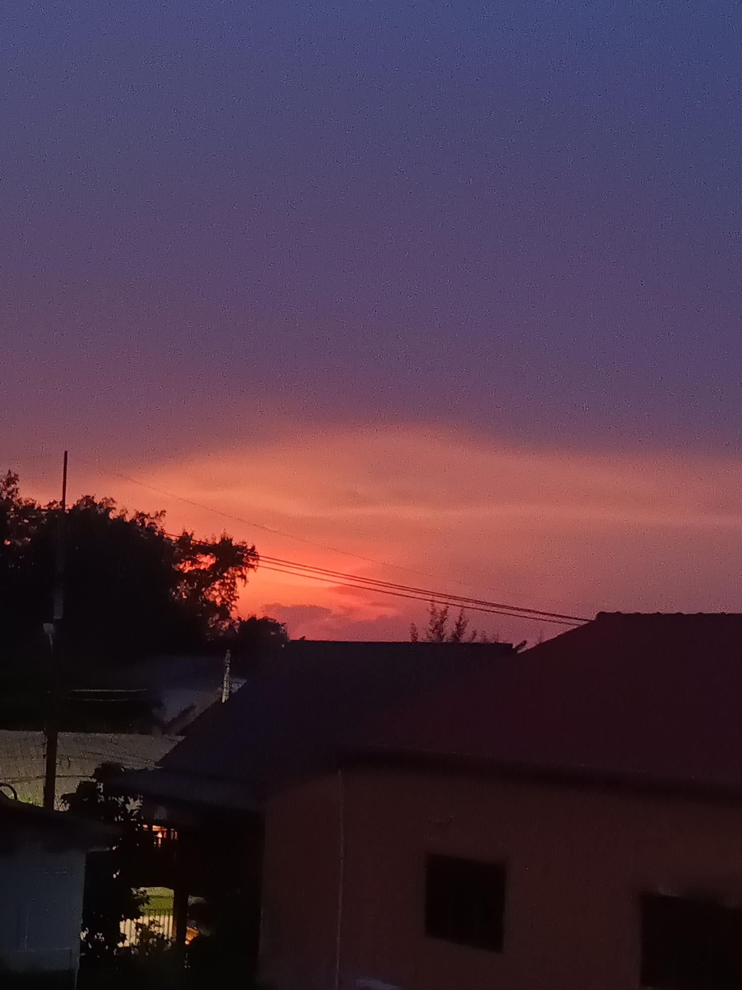 The media file displays a serene sunset or sunrise scene over Baan Chaloklum, Thailand. The sky dominates the image, showcasing a vibrant gradient of colors, transitioning from deep indigo and purple at the top to striking oranges and reds across the middle, indicating the sun's position near the horizon. Subtle cloud formations are visible, illuminated by the warm light.
In the lower portion of the image, the landscape is rendered in deep silhouette against the bright sky. Dense foliage from trees is visible on the left and center-left, with a vertical pole, possibly a utility pole, rising from the greenery. Power lines stretch horizontally across the mid-right section of the sky. Below these natural elements, the dark outlines of residential rooftops and various building structures are visible. One building on the far right features dark, rectangular window-like shapes. A structure on the far left appears slightly lighter, potentially catching some artificial light. No people or visible text are present. The image contains a noticeable amount of digital noise, particularly in the sky and shadowed areas.