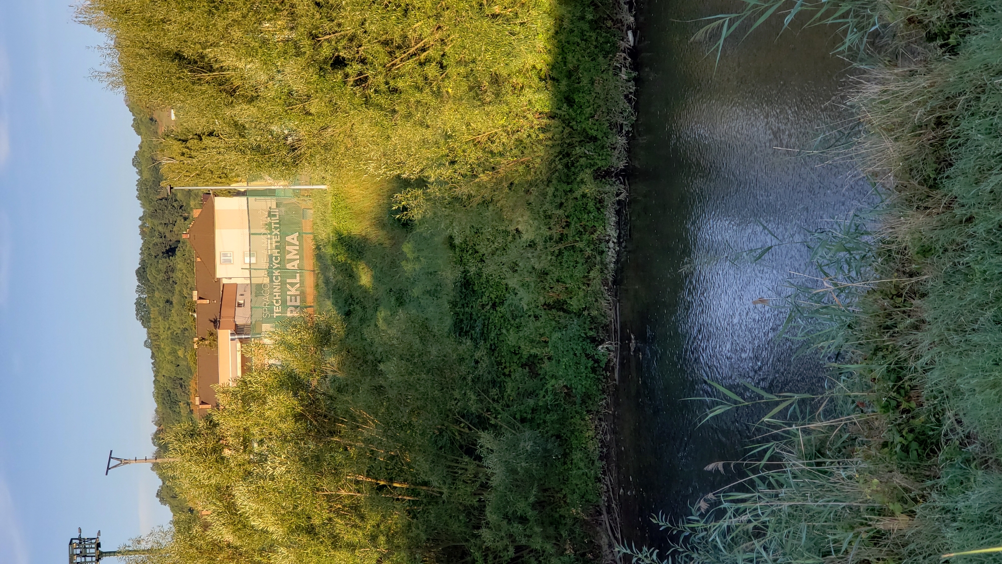 The image depicts a serene natural scene with a body of water in the foreground and buildings and lush greenery in the background. The water appears to be a river or stream, with reflections of the sky and trees on its surface. Reeds and tall grass grow along the bank, adding to the natural ambiance.
In the mid-ground, a fence with a banner is visible. The banner displays text, with the word "REKLAMA" clearly legible in large letters. Above "REKLAMA," smaller text reads "ZPRACOVANIE A MONTÁŽ TECHNICKÝCH TEXTÍLIÍ". This suggests that the fence belongs to a business involved in the processing and assembly of technical textiles, and that "reklama" is an advertisement for this business. Behind the fence, residential buildings with reddish-brown roofs are situated on a gentle slope, partially obscured by trees.
The background is dominated by a dense forest covering rolling hills, bathed in the warm light of what appears to be late afternoon or early morning sun. The sky is a clear blue with a hint of white clouds. To the left of the buildings, a tall metal pole with lights or equipment attached is visible, potentially related to sports facilities or other outdoor lighting. The overall impression is one of a peaceful setting where nature and human settlement coexist.
This scene is located in Prešov, Slovakia, specifically in the area of the local tennis club (Tenisový areál Prešov), which is known to have a fence with similar advertising banners. The presence of the tennis court surface is hinted at by the texture visible behind the fence in the cropped images.