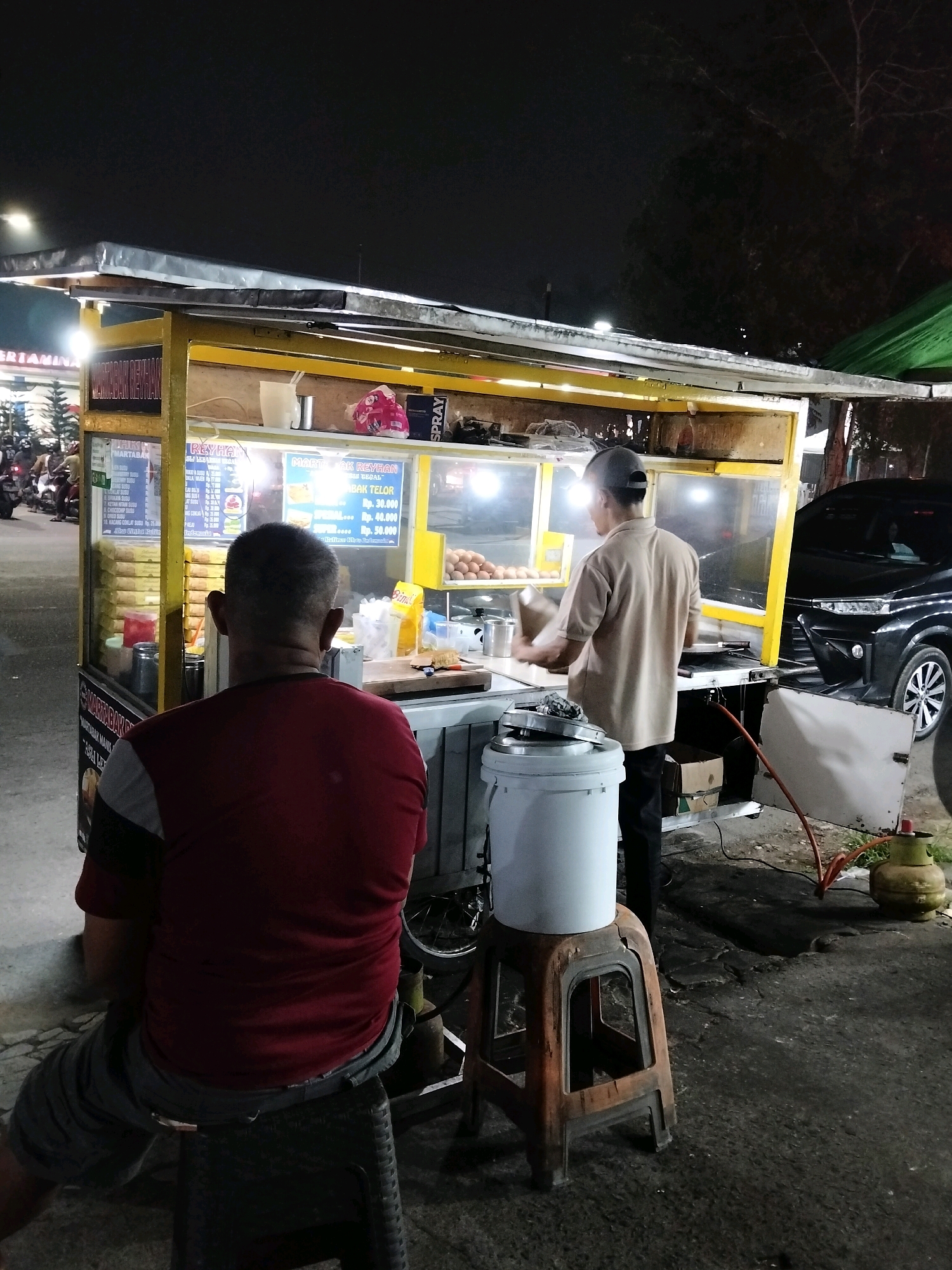 The image depicts an outdoor food stall at night, likely in Pontianak, Indonesia, evidenced by the Indonesian language on the menu boards. A man wearing a red shirt and gray shorts is seated on a stool, facing away from the camera, towards the stall. He appears to be a customer. Behind him, a food vendor, wearing a light-colored shirt and a baseball cap, is actively preparing food at the stall. The stall is illuminated by bright lights, showcasing a display of food items and ingredients, including eggs. A black car is parked to the right of the stall, and a green gas cylinder is visible on the ground near the stall. The overall environment suggests a busy street food scene.